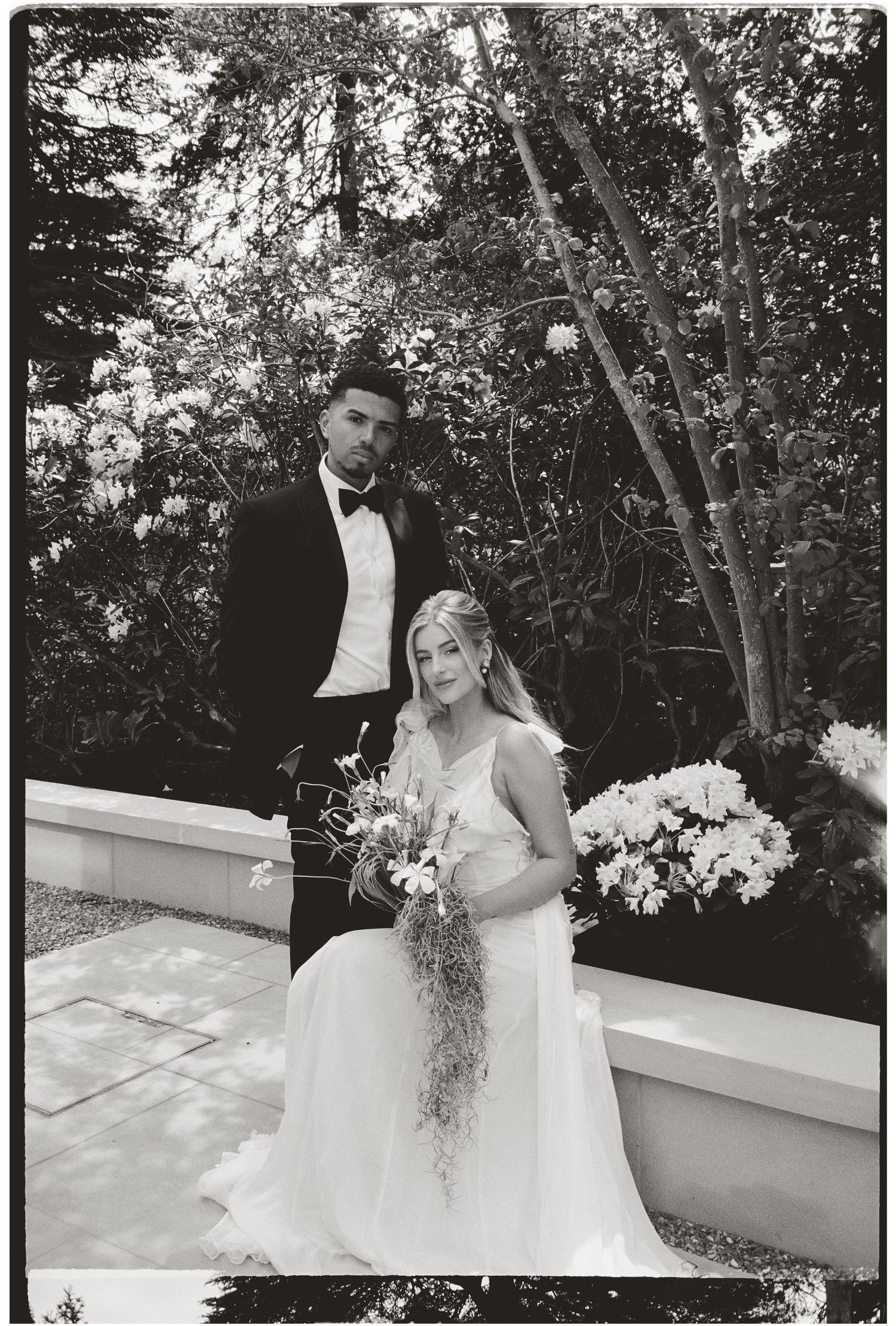 Black and white wedding photo of bride in white gown holding bouquet with groom in tuxedo amid lush garden backdrop.