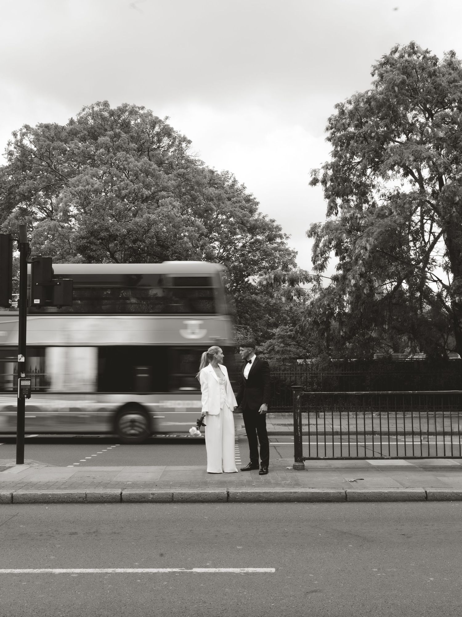 Black and white photo of a bride and groom standing at a London bus stop with a double-decker bus passing by.