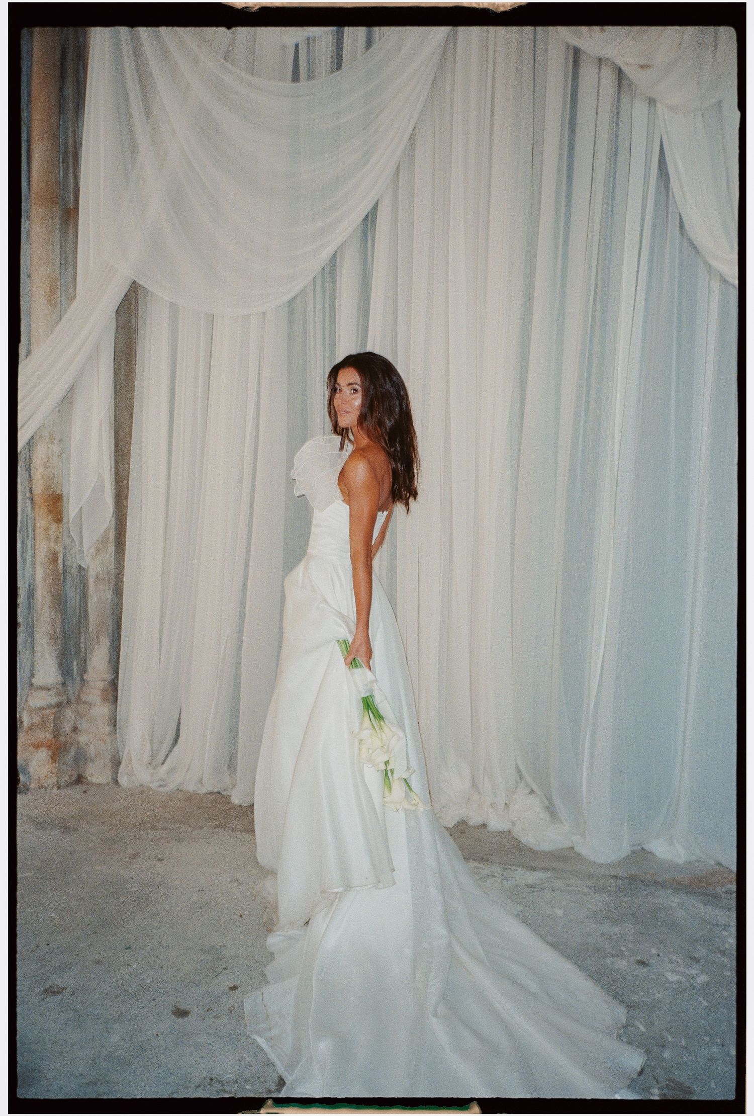 Bride in elegant backless white gown against light fabric backdrop.