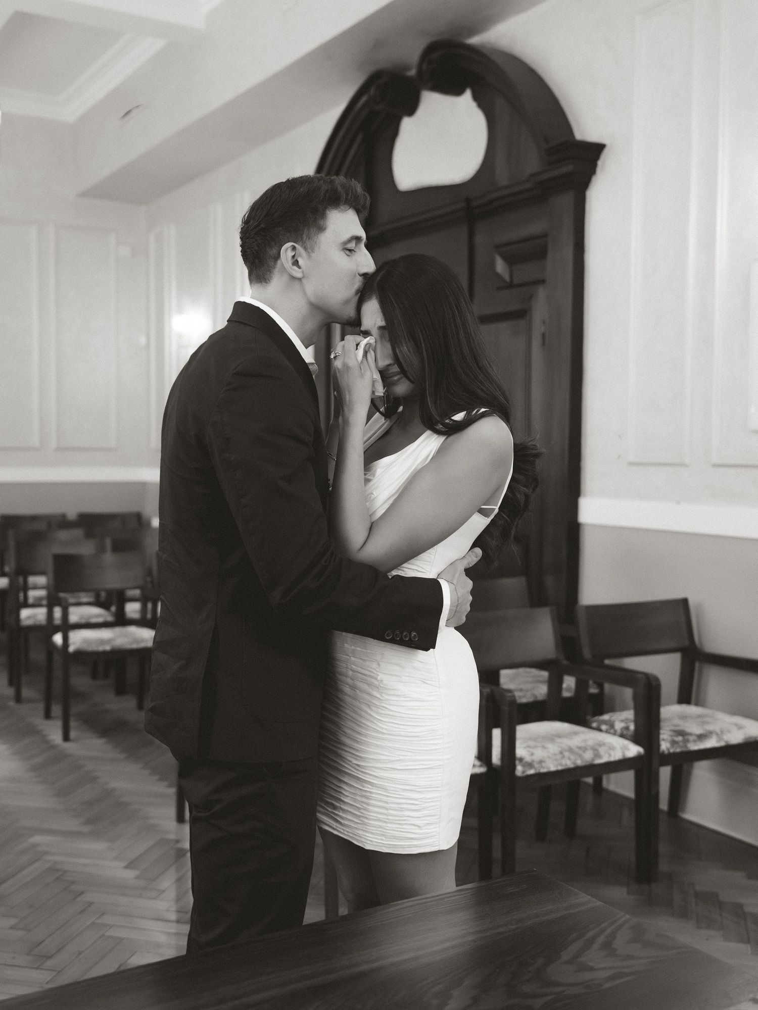 Black and white photo of an elegant couple embracing in a formal room, man in suit and woman in white dress.