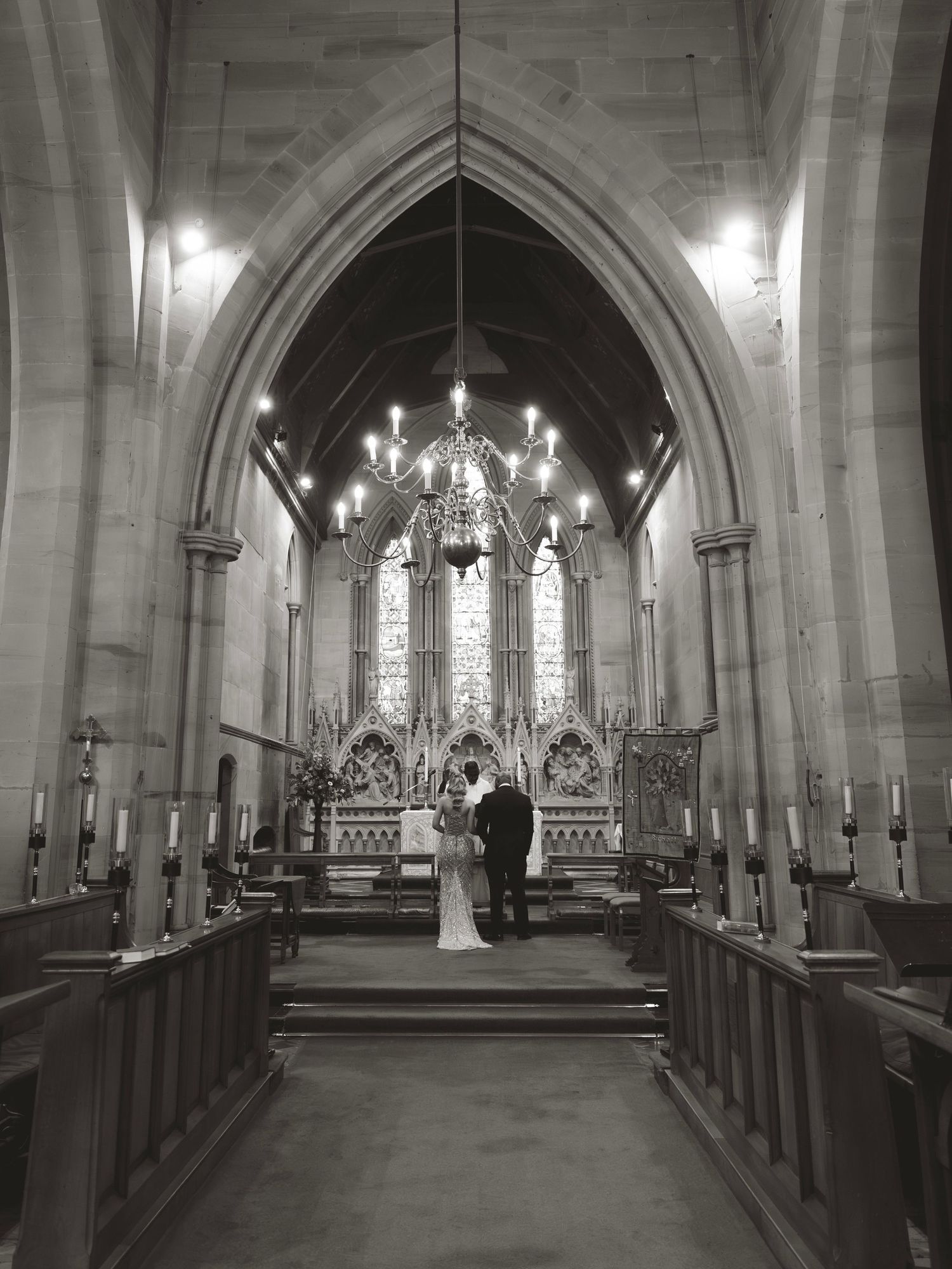 Black and white photo of a couple exchanging vows at a church altar with gothic arches and chandelier lighting.