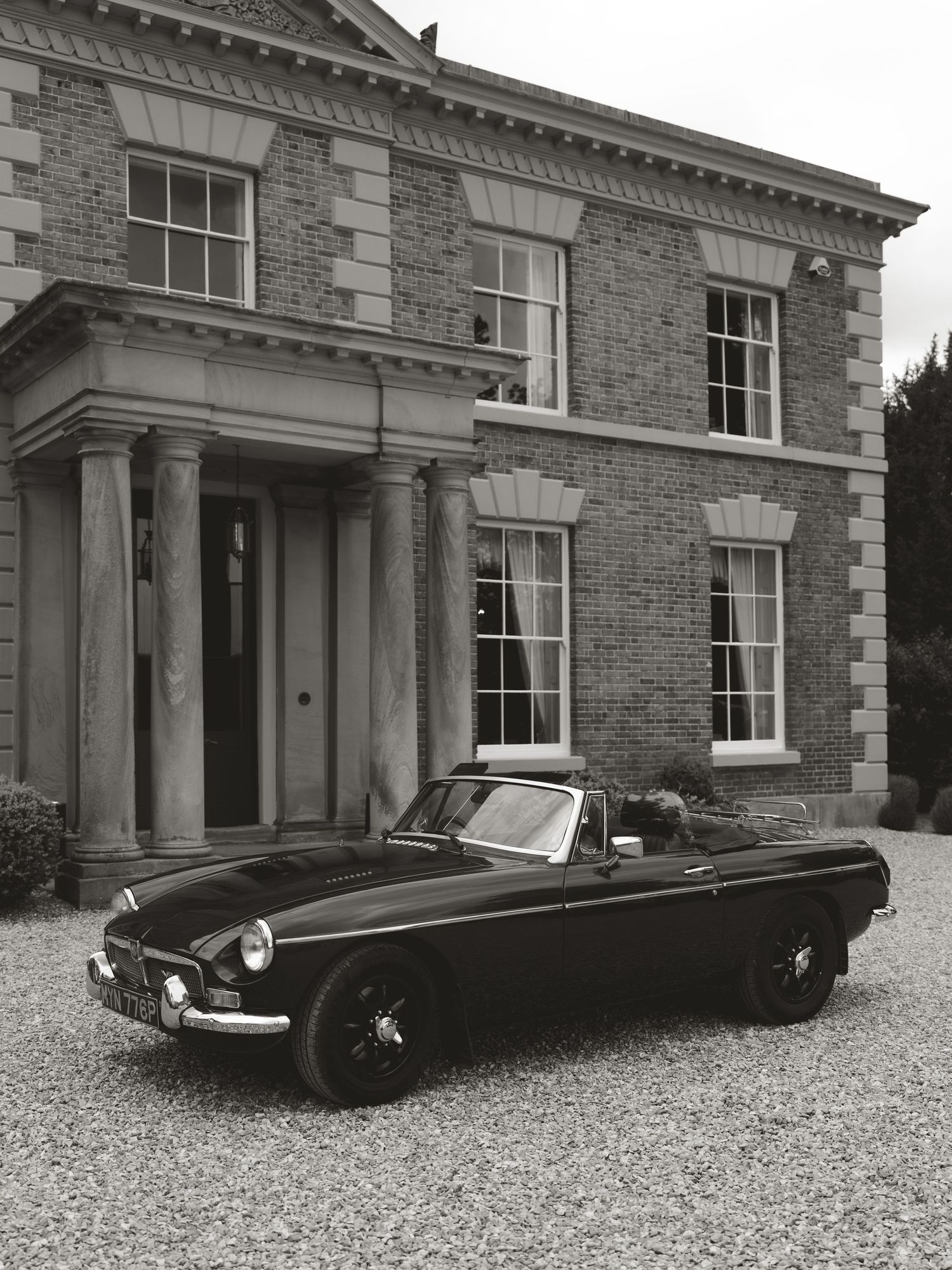 Vintage MGB roadster convertible parked on gravel driveway before a classic Georgian brick manor house, black and white photo.