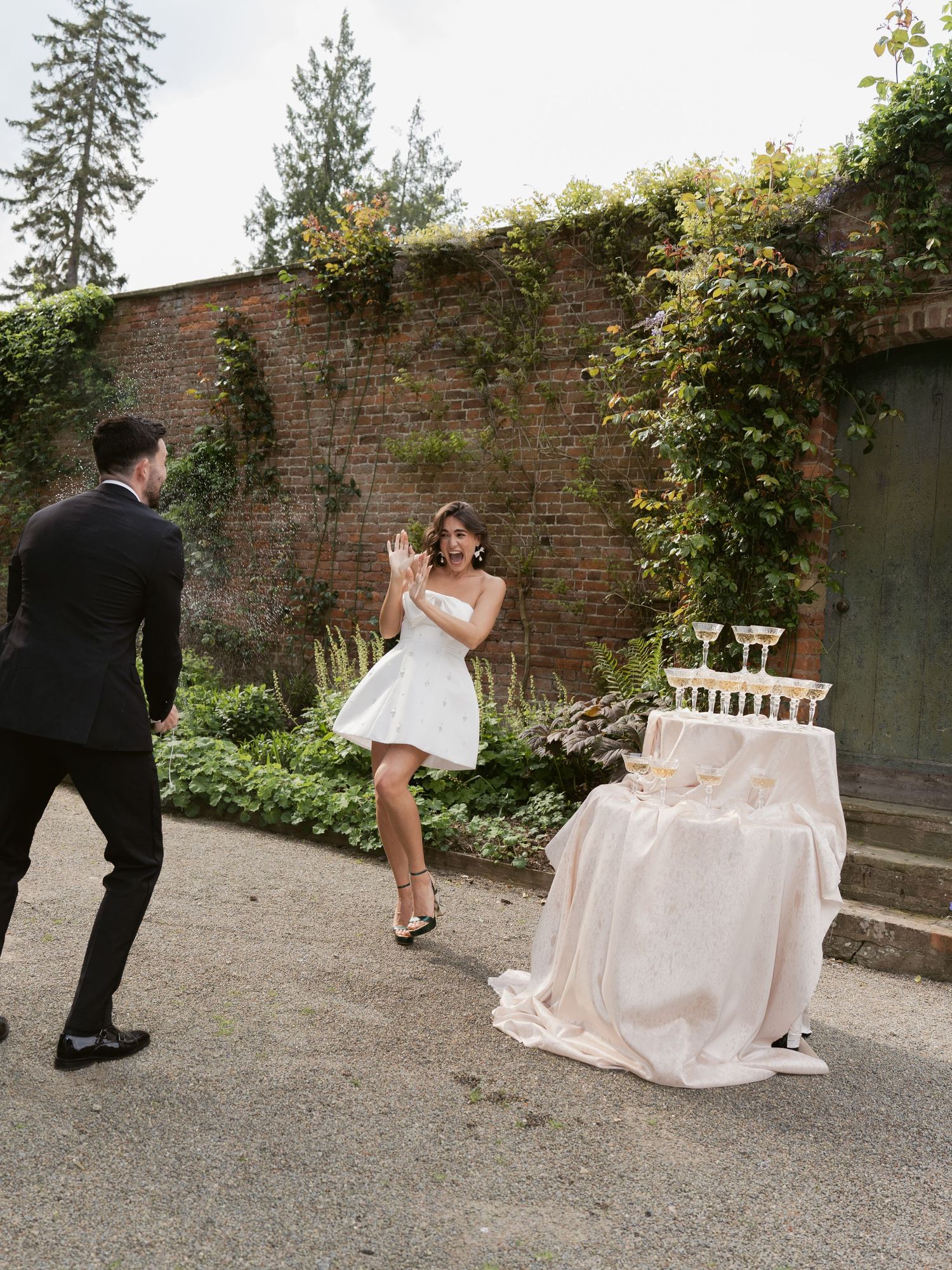 Bride in white mini dress laughing with groom near champagne tower at outdoor wedding reception by brick wall.