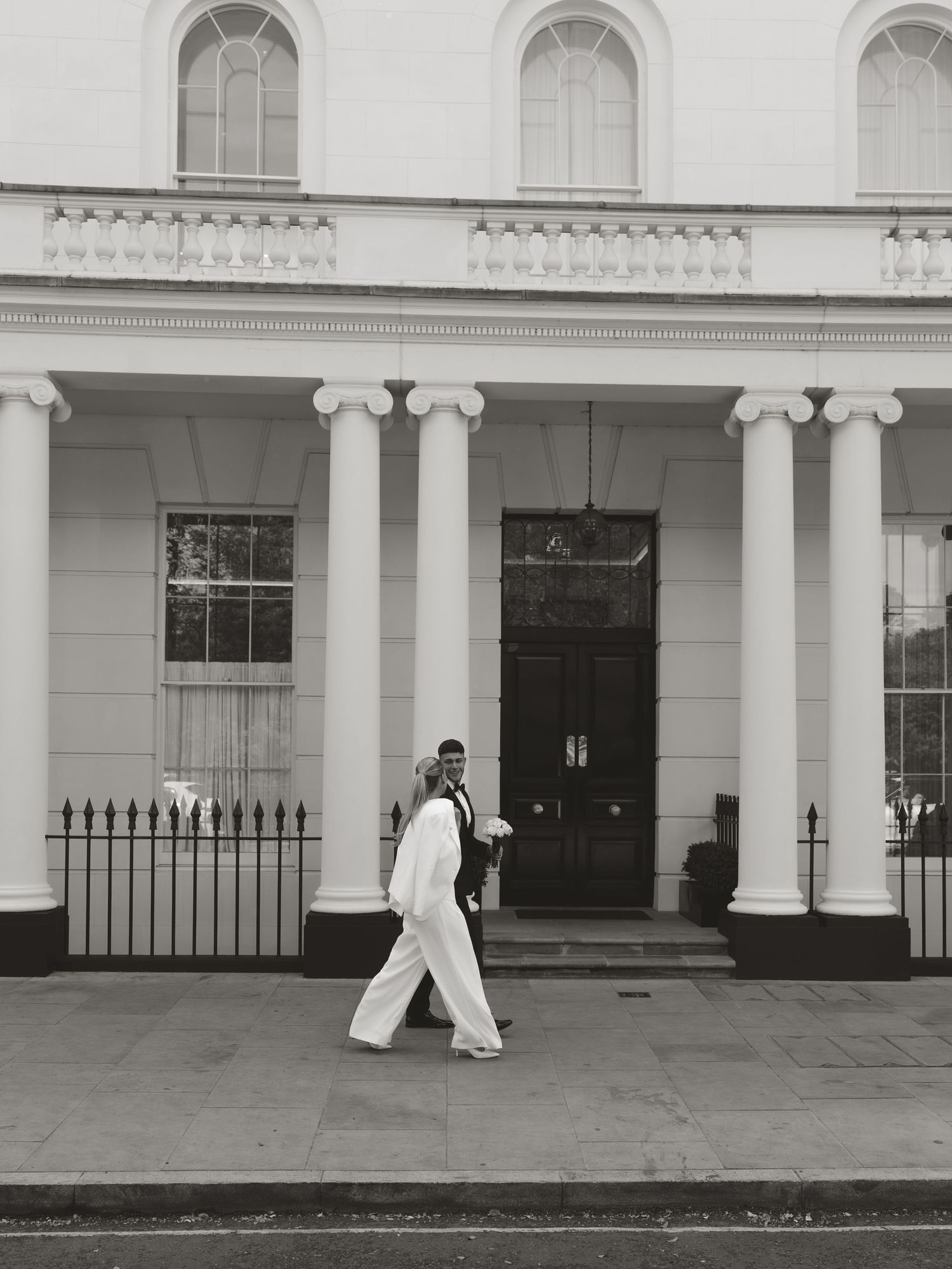 Black and white photo of a couple in wedding attire walking past a grand white neoclassical building with columns.