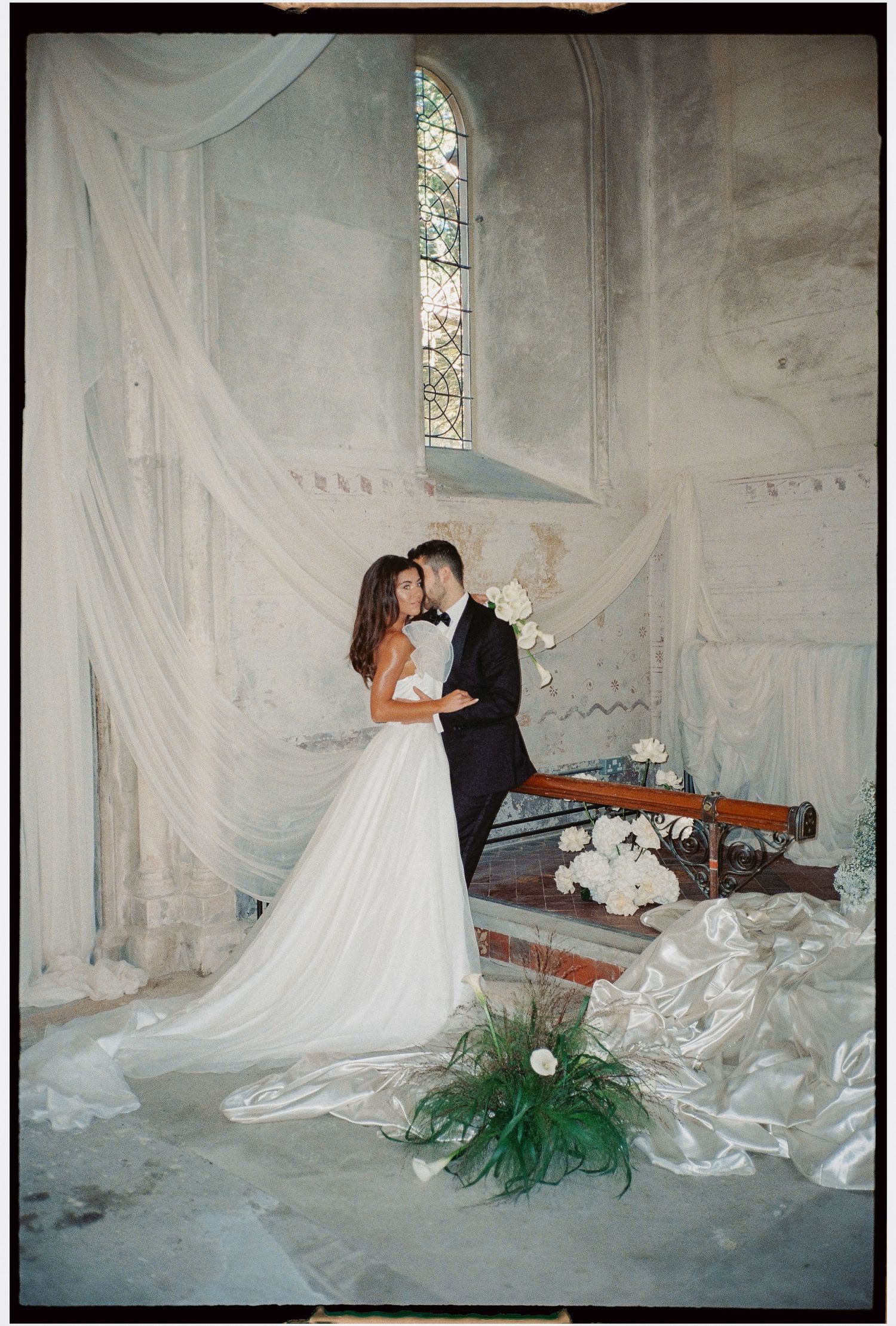 Wedding couple in romantic pose on vintage bench with white fabric draping.