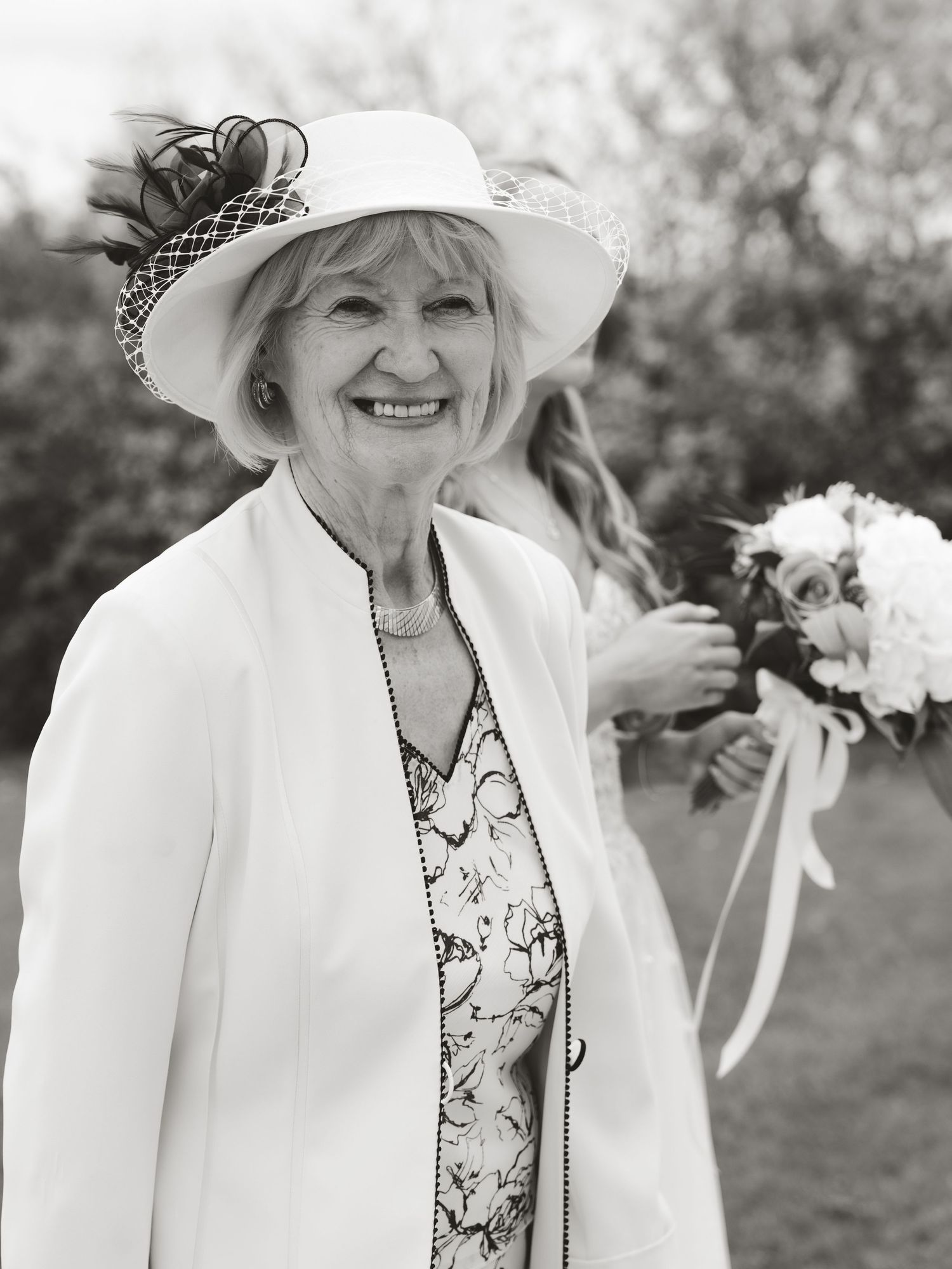 Elegant elderly woman smiling in white hat and jacket at outdoor wedding ceremony with bouquet in background.