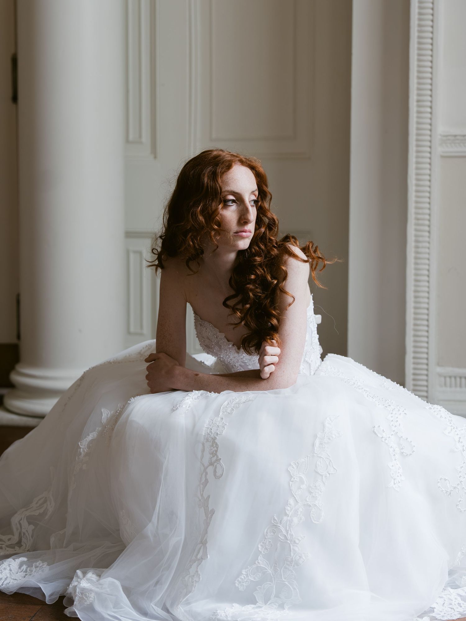 A bride in a white lace ball gown sits gracefully between white columns with flowing red curly hair.