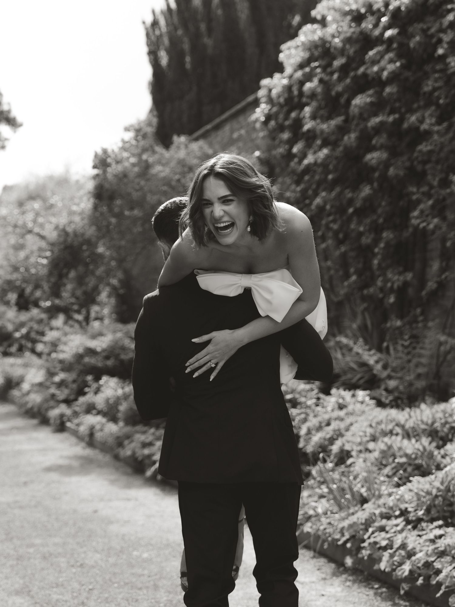 Joyful woman laughing while getting a piggyback ride from a man in a suit in a lush garden, black and white photo.