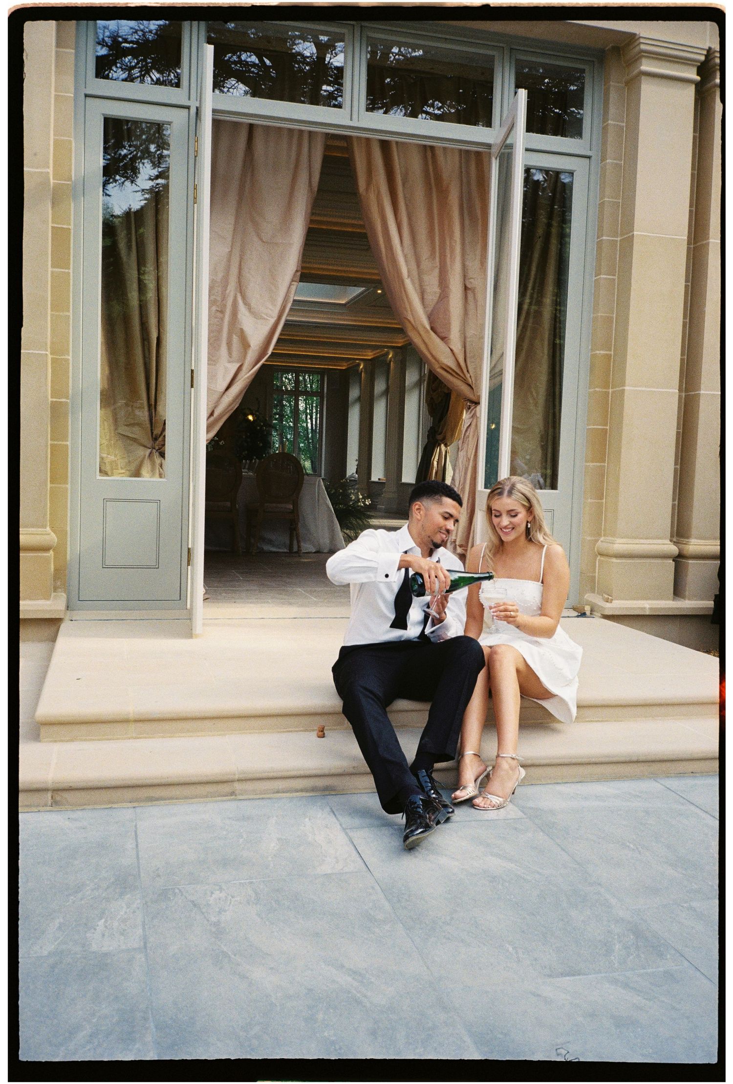 Couple in wedding attire sitting on steps of elegant venue, pouring champagne together on their wedding day.