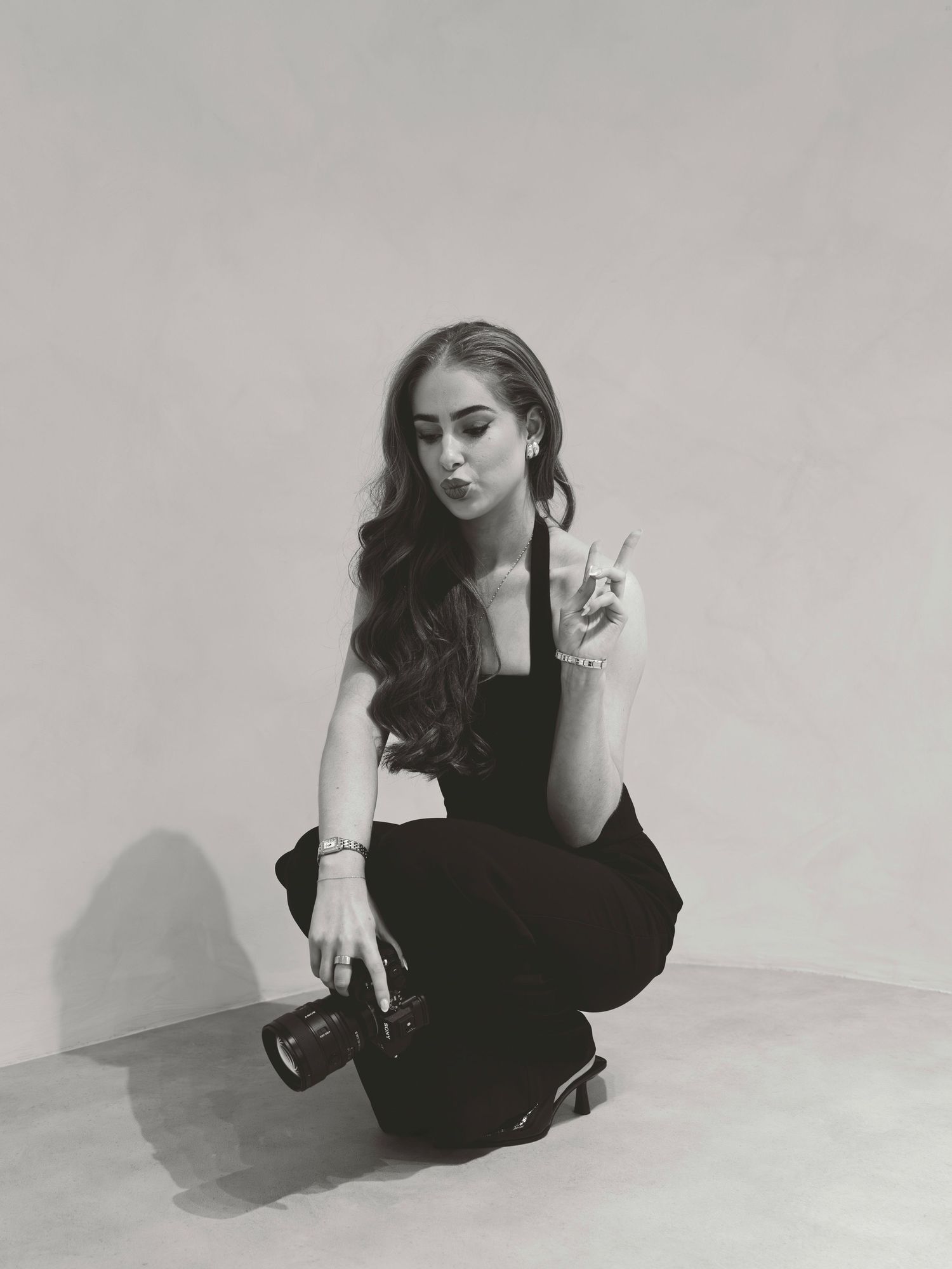 Black and white photo of a young woman crouching, holding a camera, making a peace sign, wearing a black outfit.