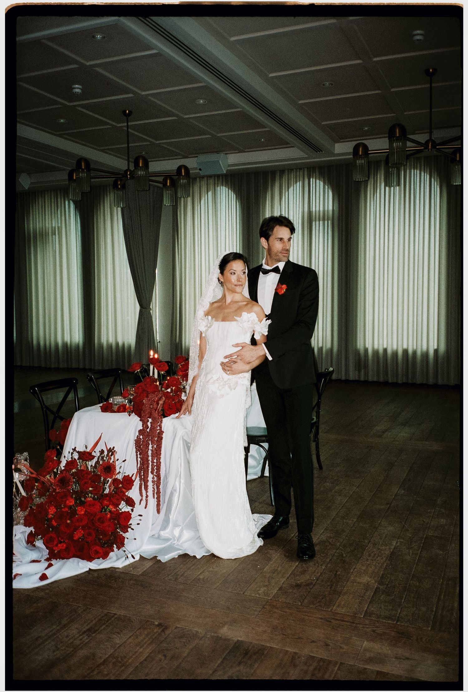 A couple shares an intimate dance moment on a wooden floor with vibrant red floral arrangements in the background.