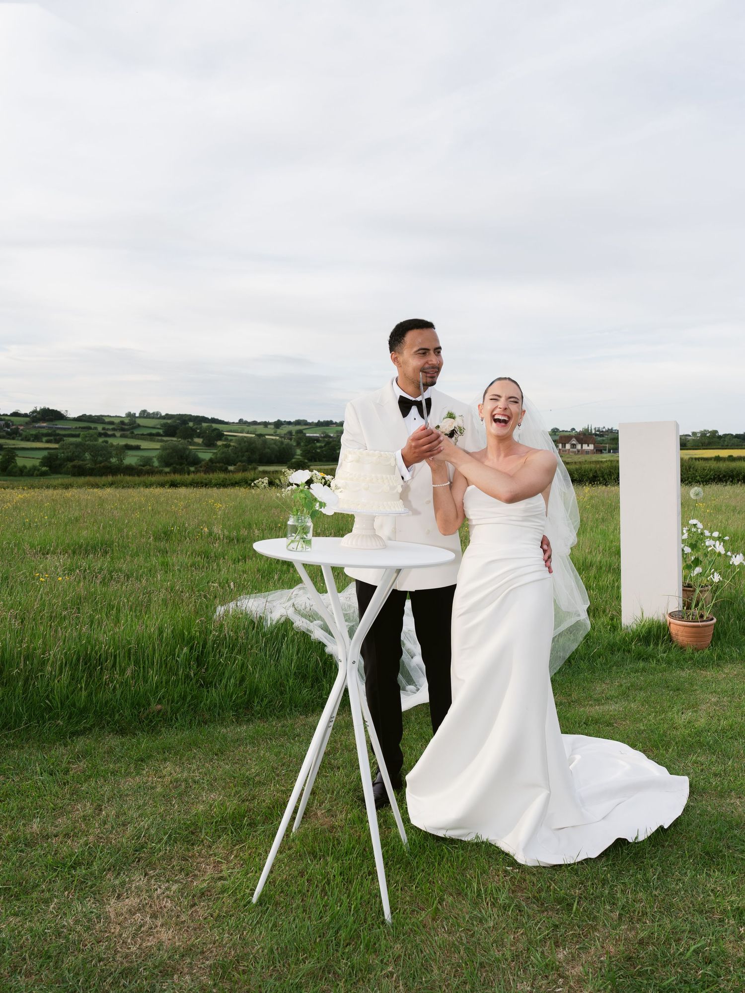 Bride and groom laughing outdoors at wedding ceremony in green field, wearing white gown and tuxedo.