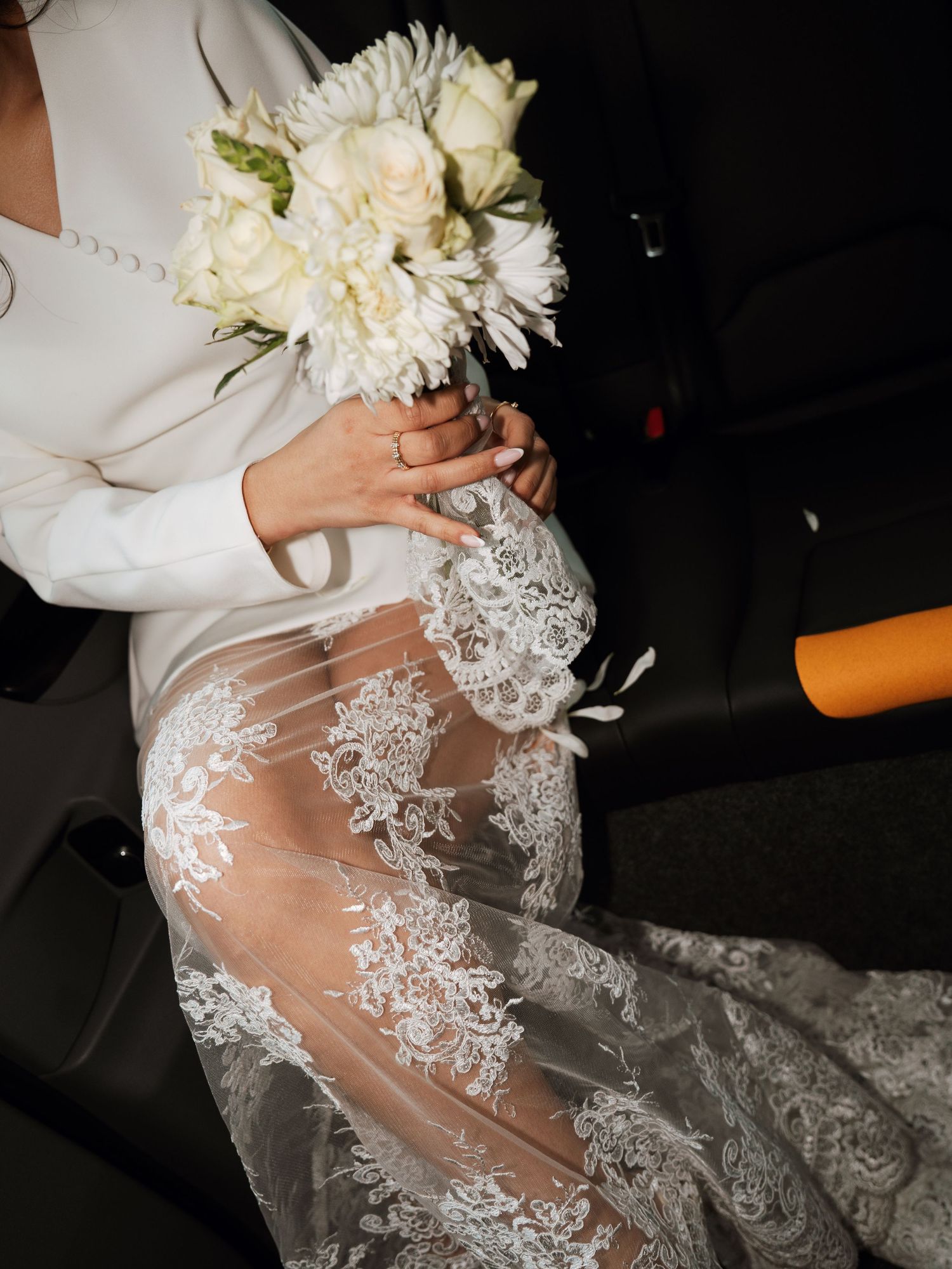 Close-up details of intricate lace wedding dress and white bouquet in car interior.