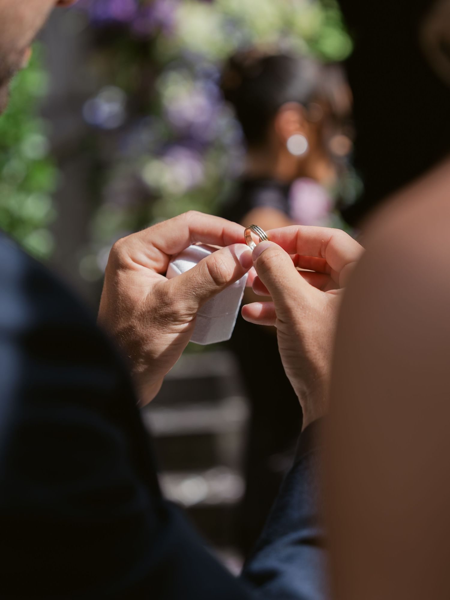 Close-up of hands holding a wedding ring over a white box during an outdoor ceremony with purple flowers.