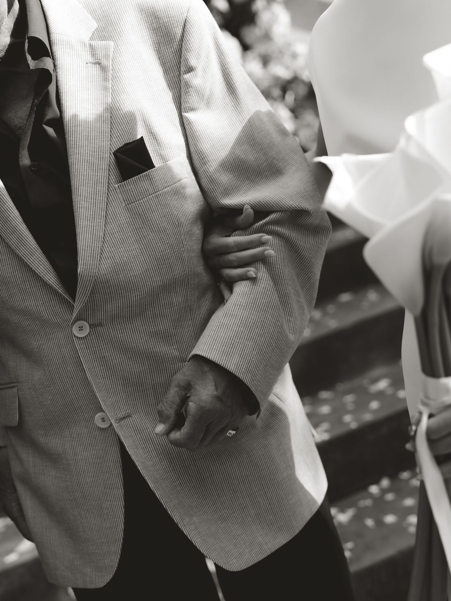 Black and white photo of a groom in a light gray suit holding hands with bride in white dress at outdoor wedding.
