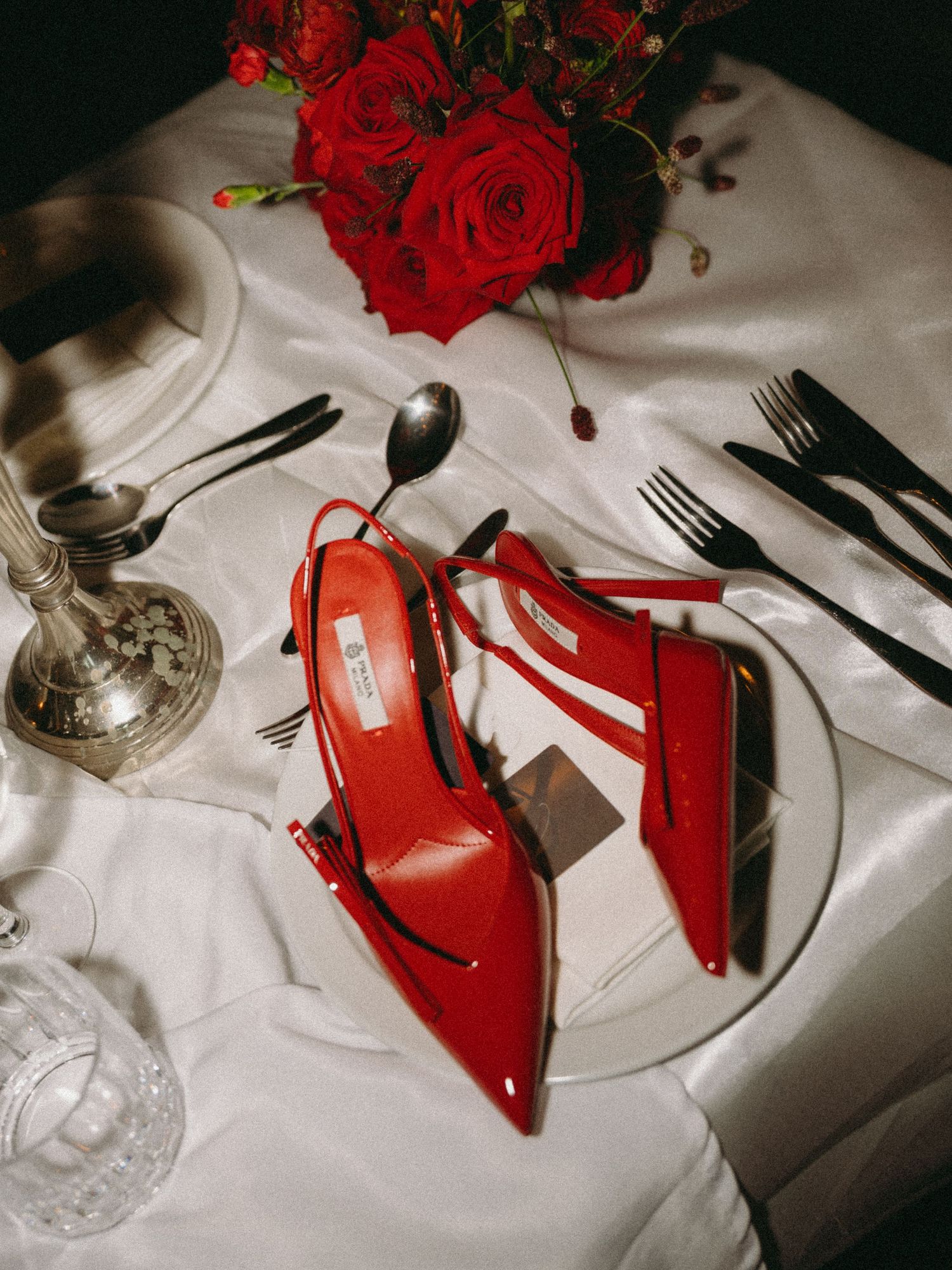 Red high heel shoes and roses create an elegant table setting with wine glasses and silverware against white linens.
