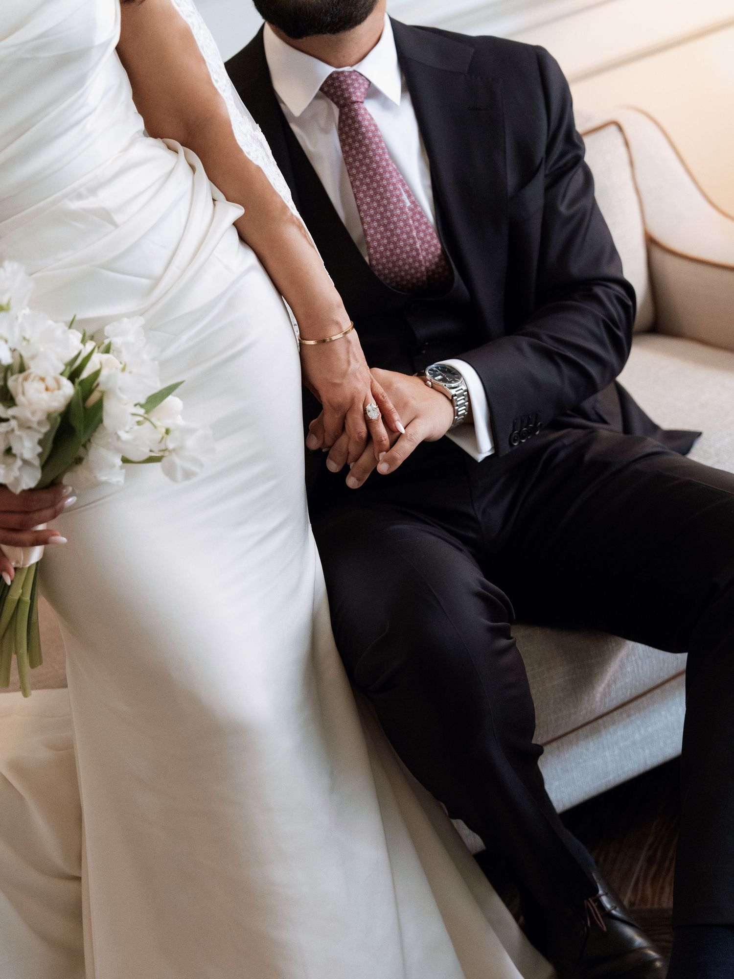 Bride and groom sit together on light colored couch holding hands with white bouquet and formal wedding attire.