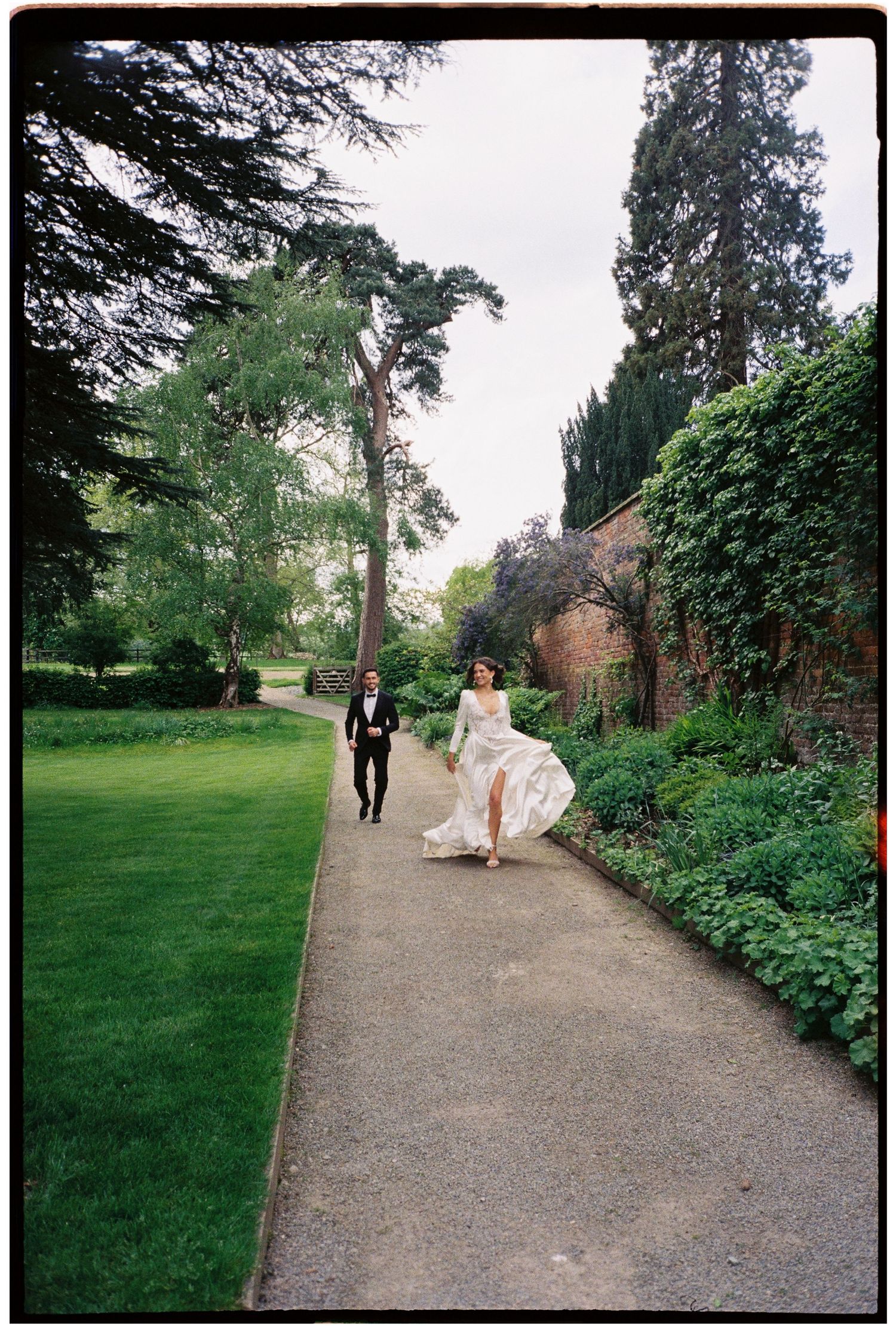 Bride in flowing white gown and groom in tuxedo walking along garden path surrounded by lush greenery.