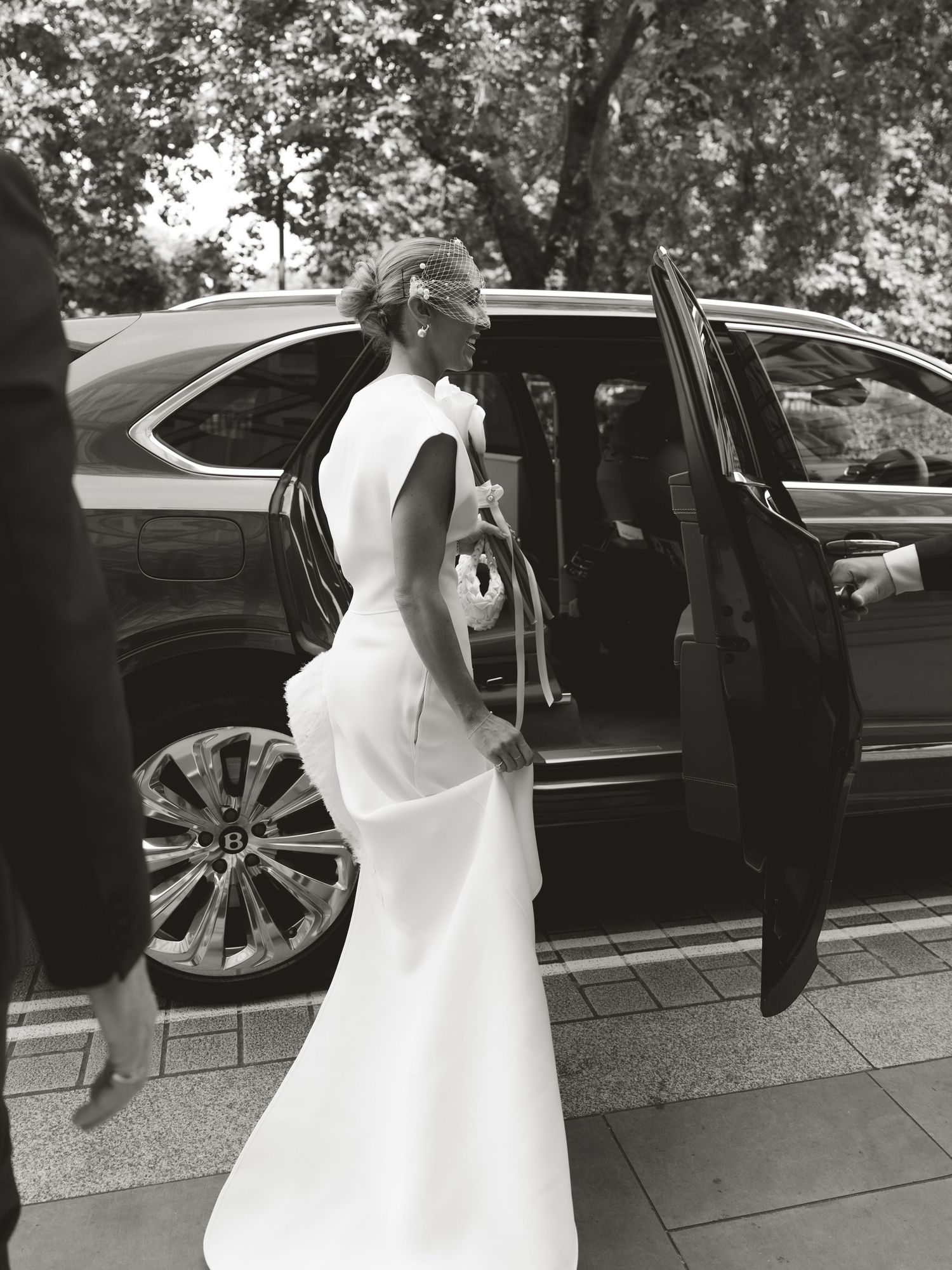 Black and white photo of a bride in a white gown entering a luxury car on her wedding day.