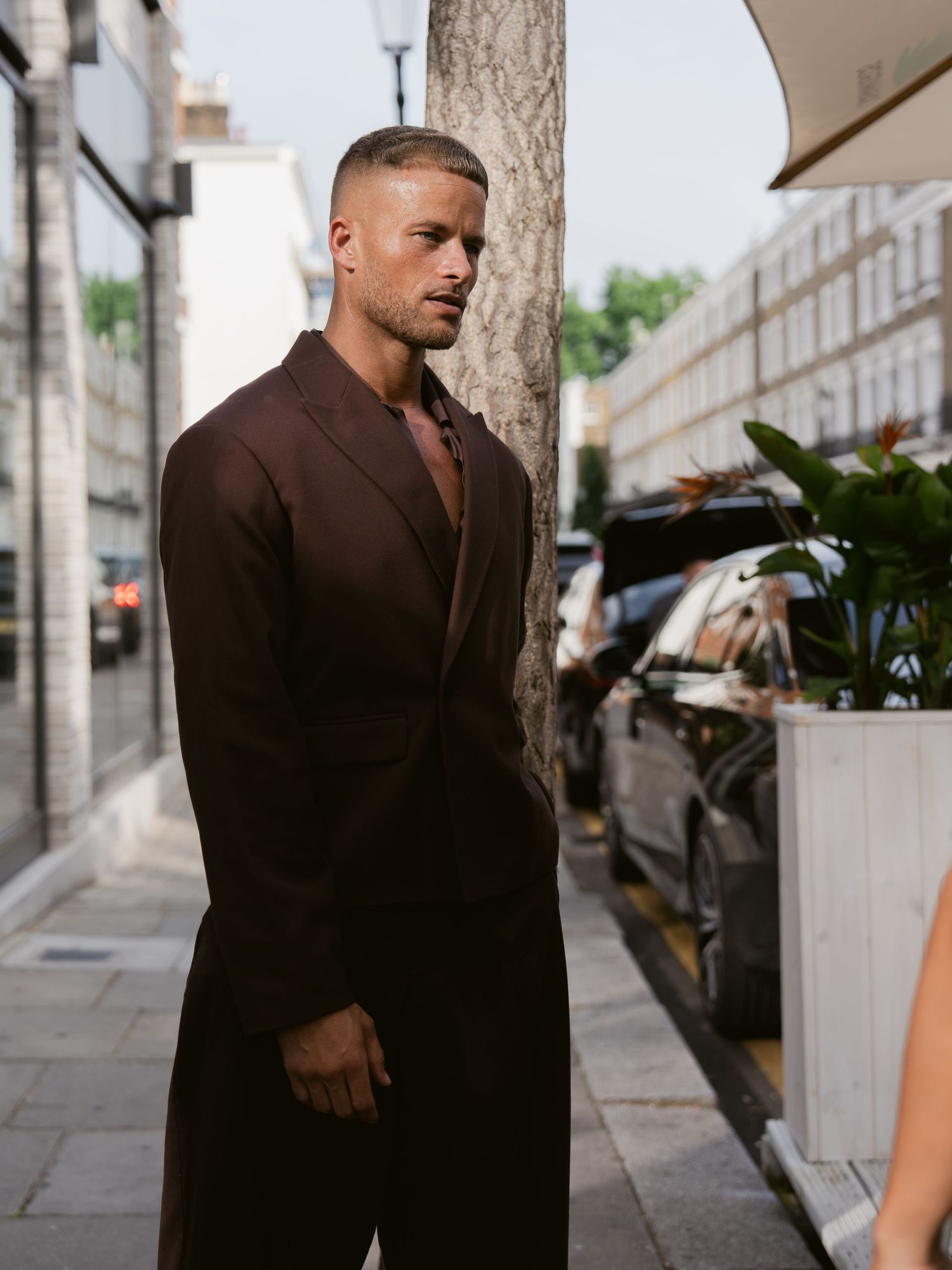 Man in brown oversized suit standing on urban sidewalk near tree, street style fashion photography.