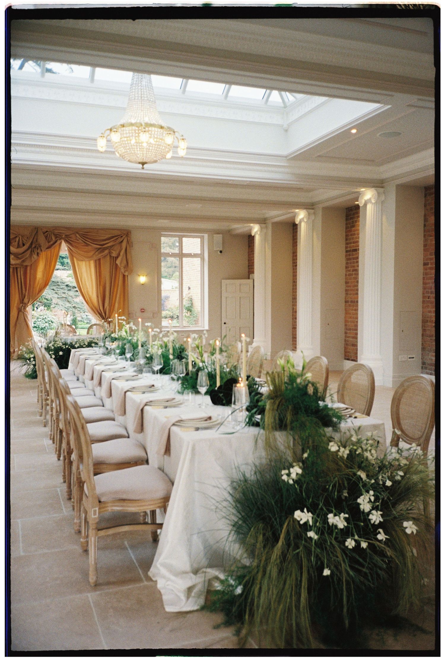 Elegant wedding reception with long table, lush greenery, candles, white linens, chandelier, and gold drapes in a bright conservatory.