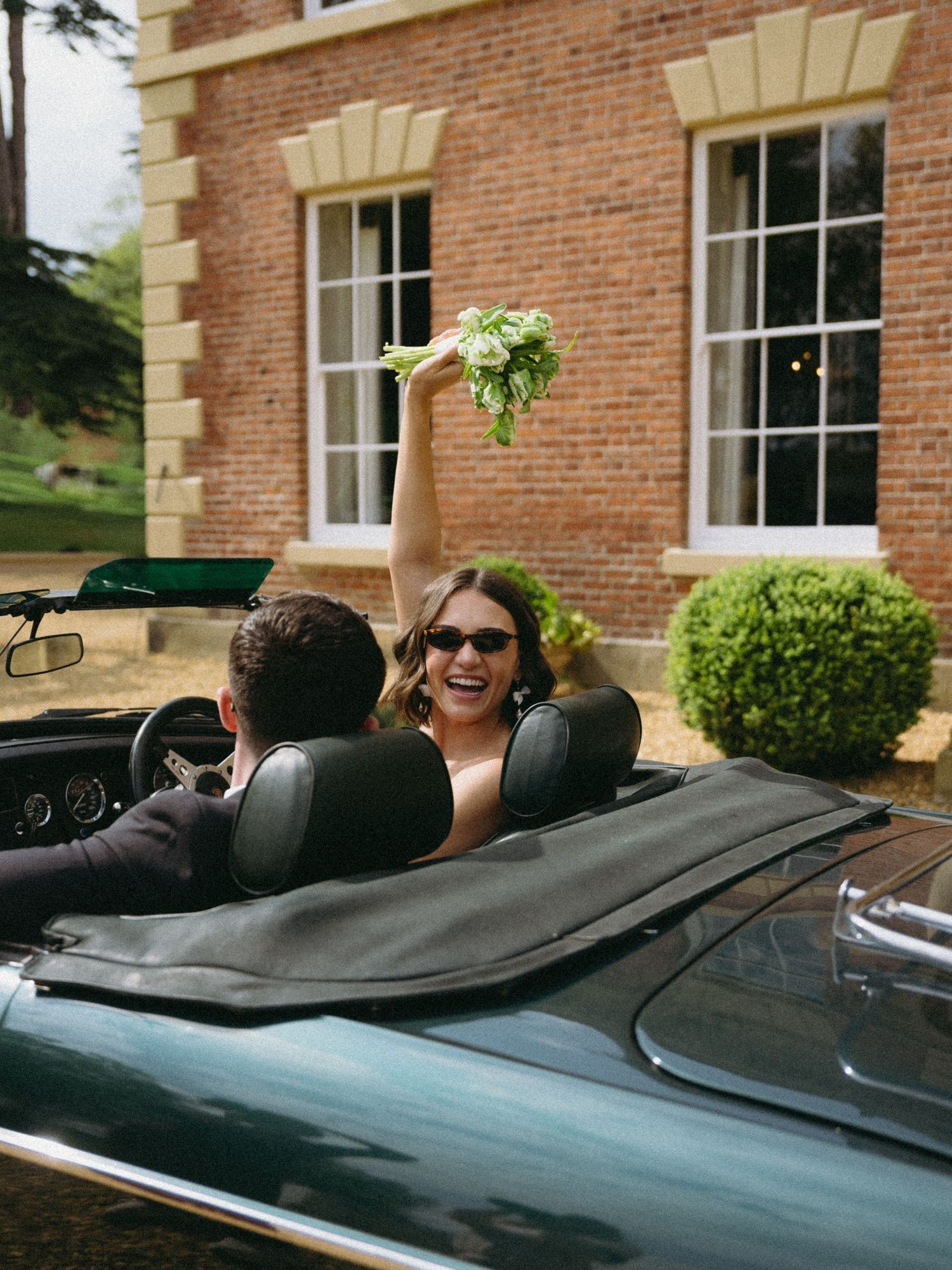 Bride in sunglasses raises bouquet from vintage green convertible car outside brick manor house.