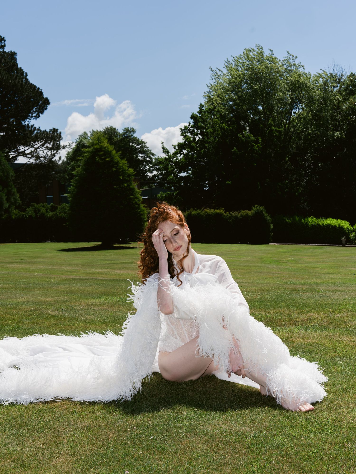 A woman with red hair sits on green grass wearing a white feathered gown, posing in a sunny garden with trimmed hedges.