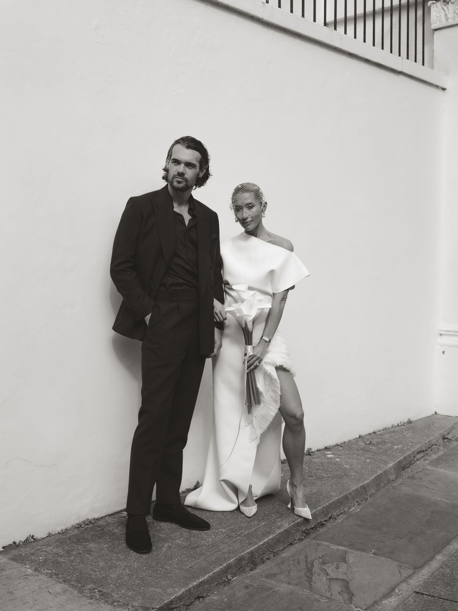 Black and white photo of a couple posing by a white wall, man in dark suit, woman in elegant white gown.