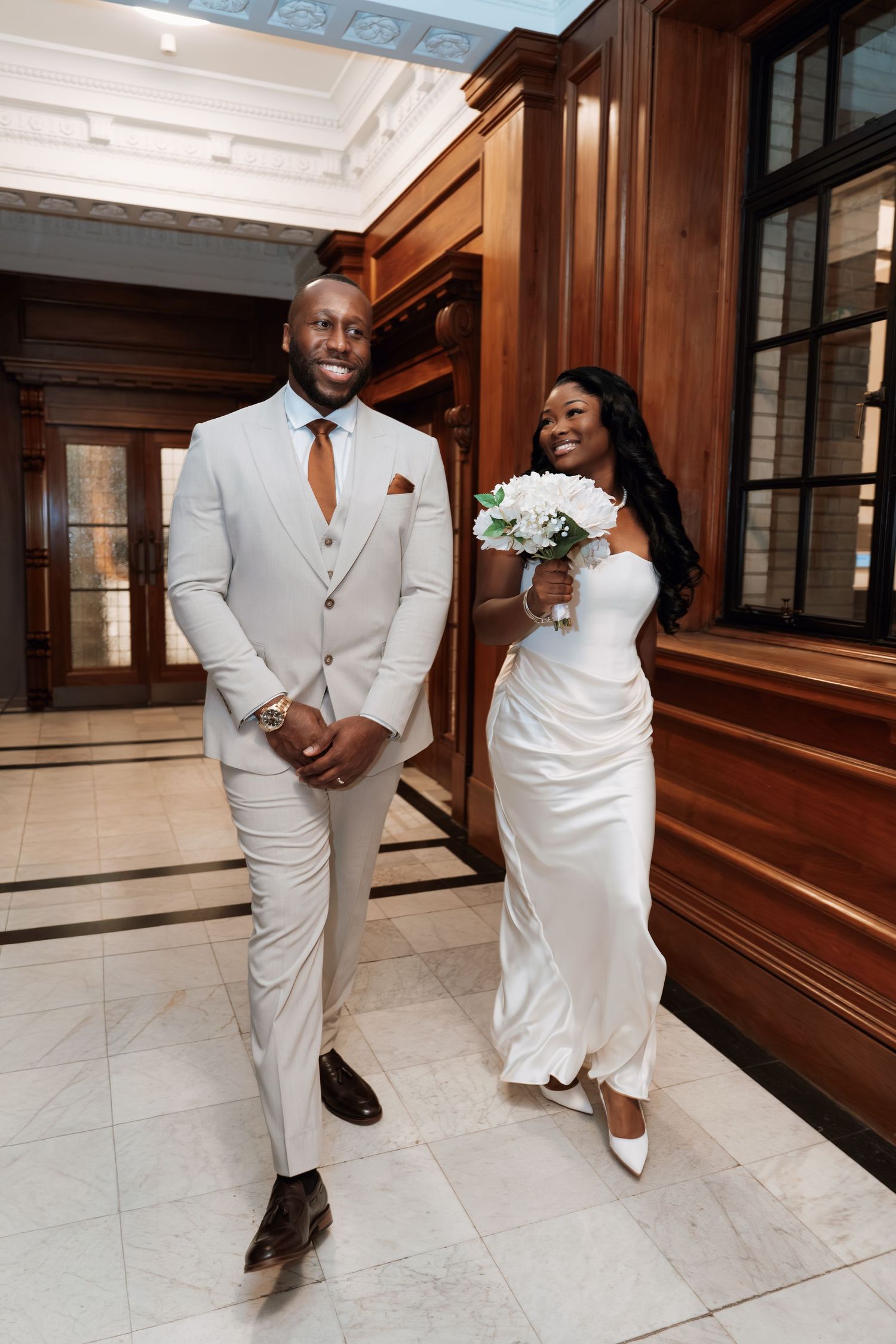 Couple in matching white formal attire walking through an elegant marble hallway.