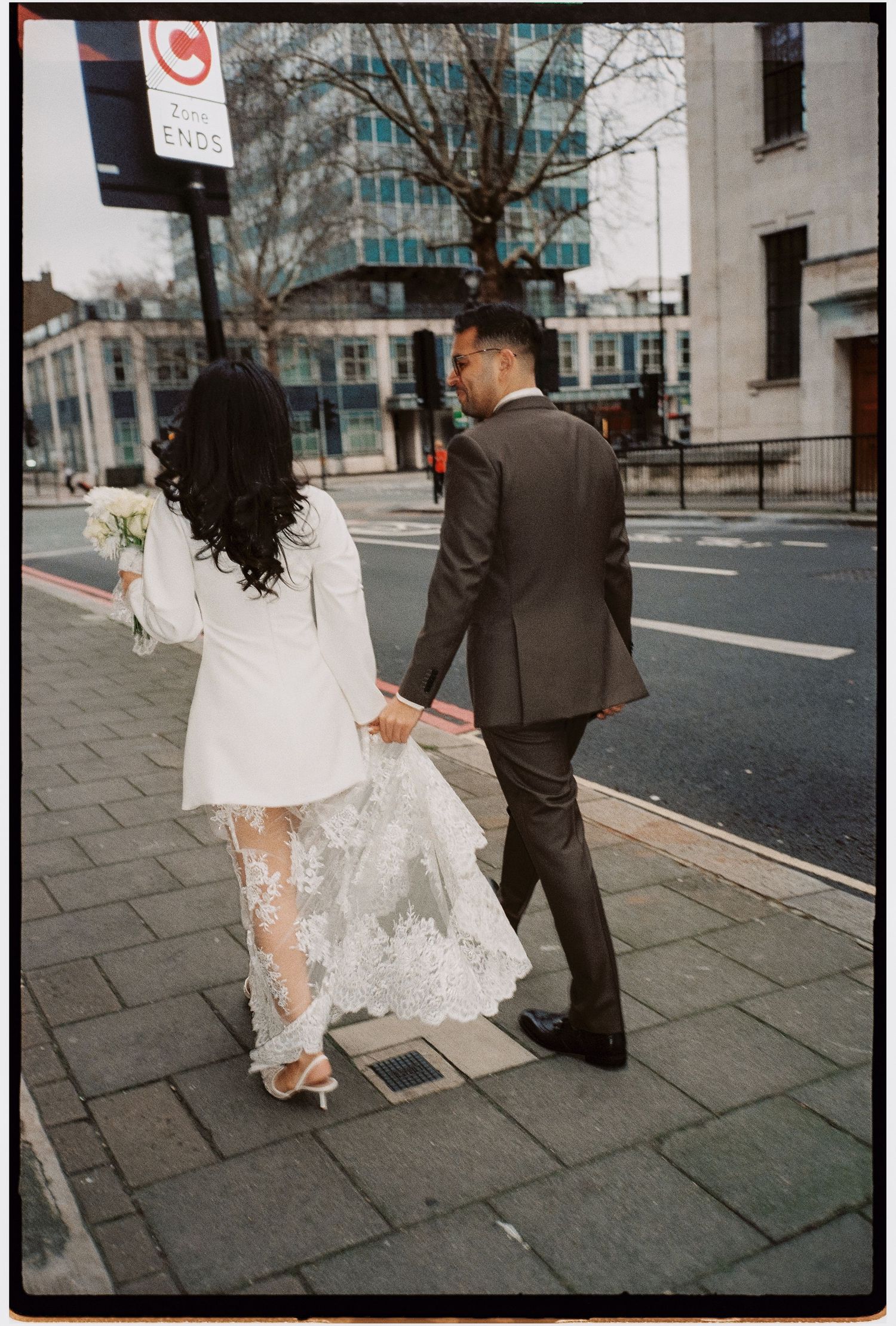 A couple walking down a city street, with the bride wearing a sheer lace-detailed dress.
