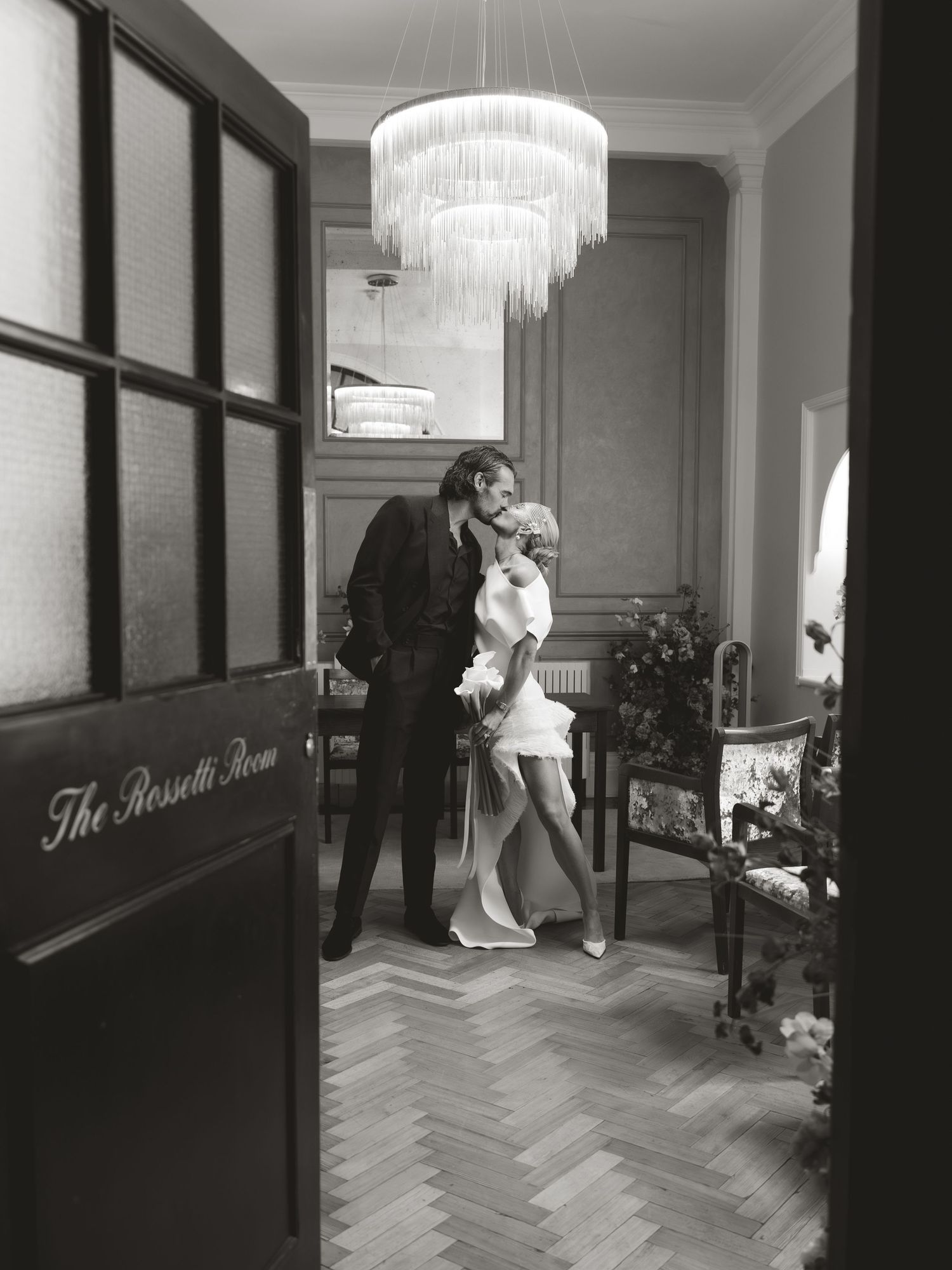 Black and white photo of a couple kissing in The Rossetti Room under a chandelier, bride in short white dress.