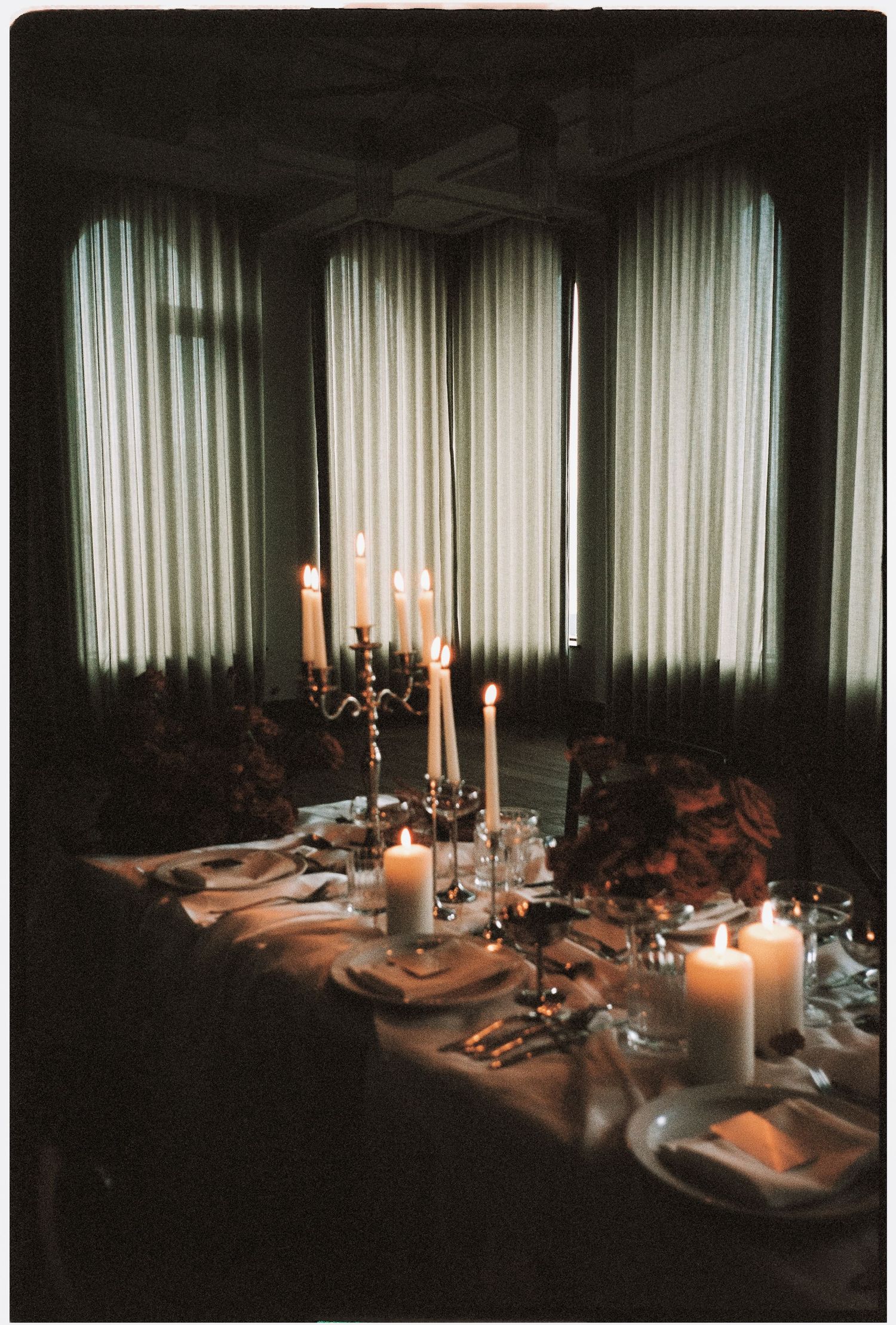 A moody dining table setting with glowing candles and white linens against a backdrop of sheer curtains in dim lighting.