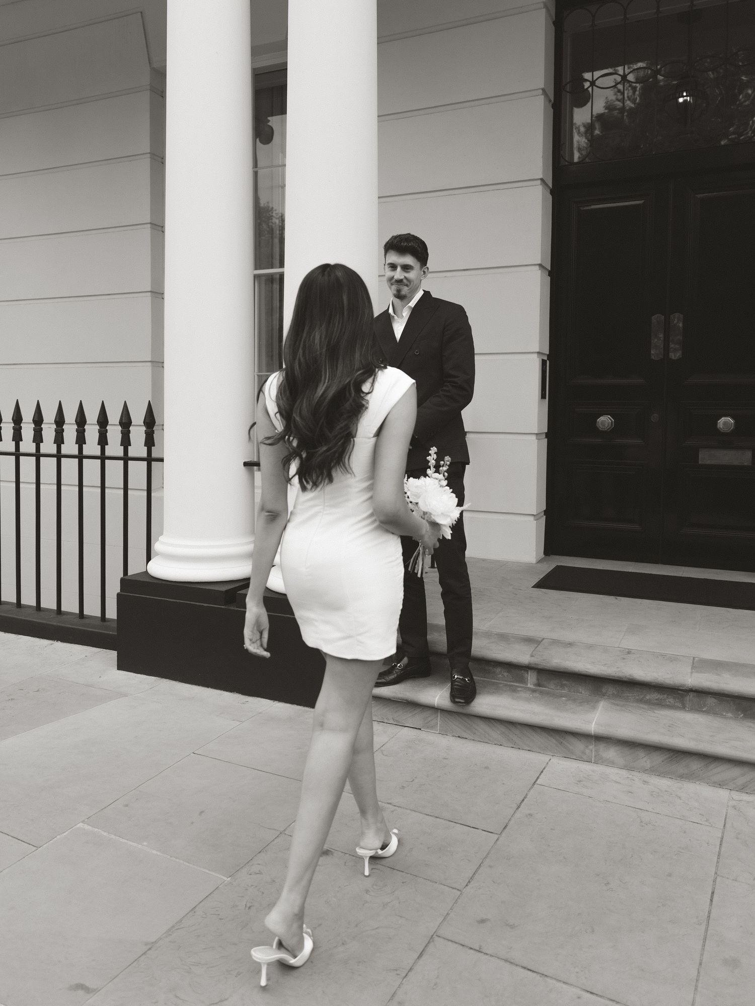 Black and white photo of a bride in a white mini dress holding flowers, walking toward a groom in a suit on building steps.