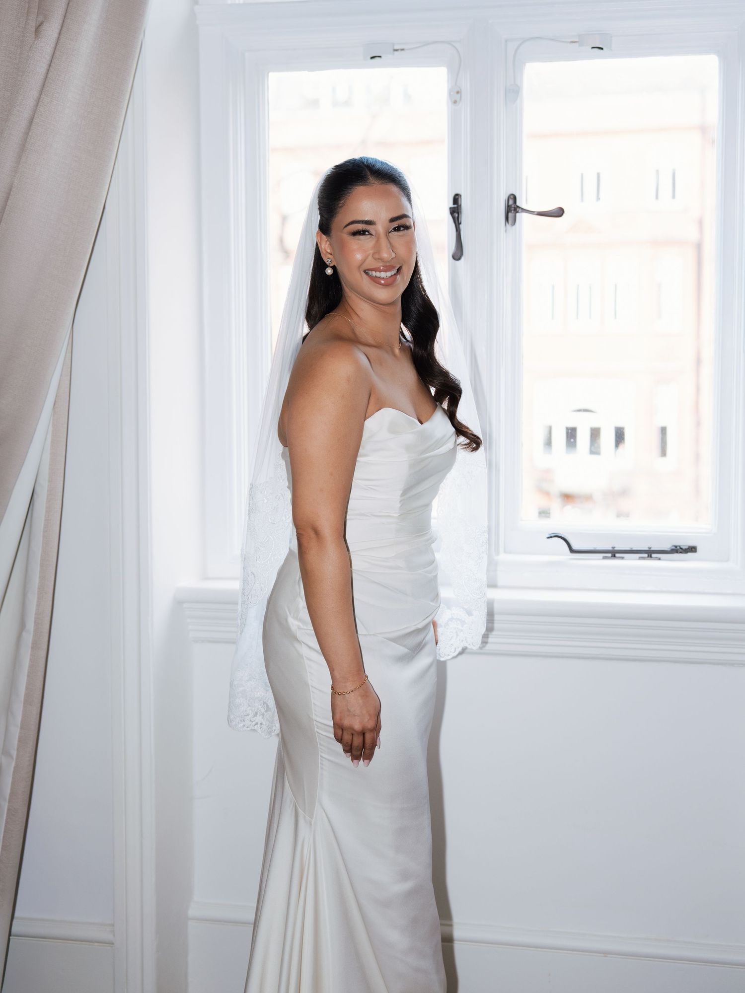 A bride in a white wedding dress stands in a bright sunlit room with white walls and windows.
