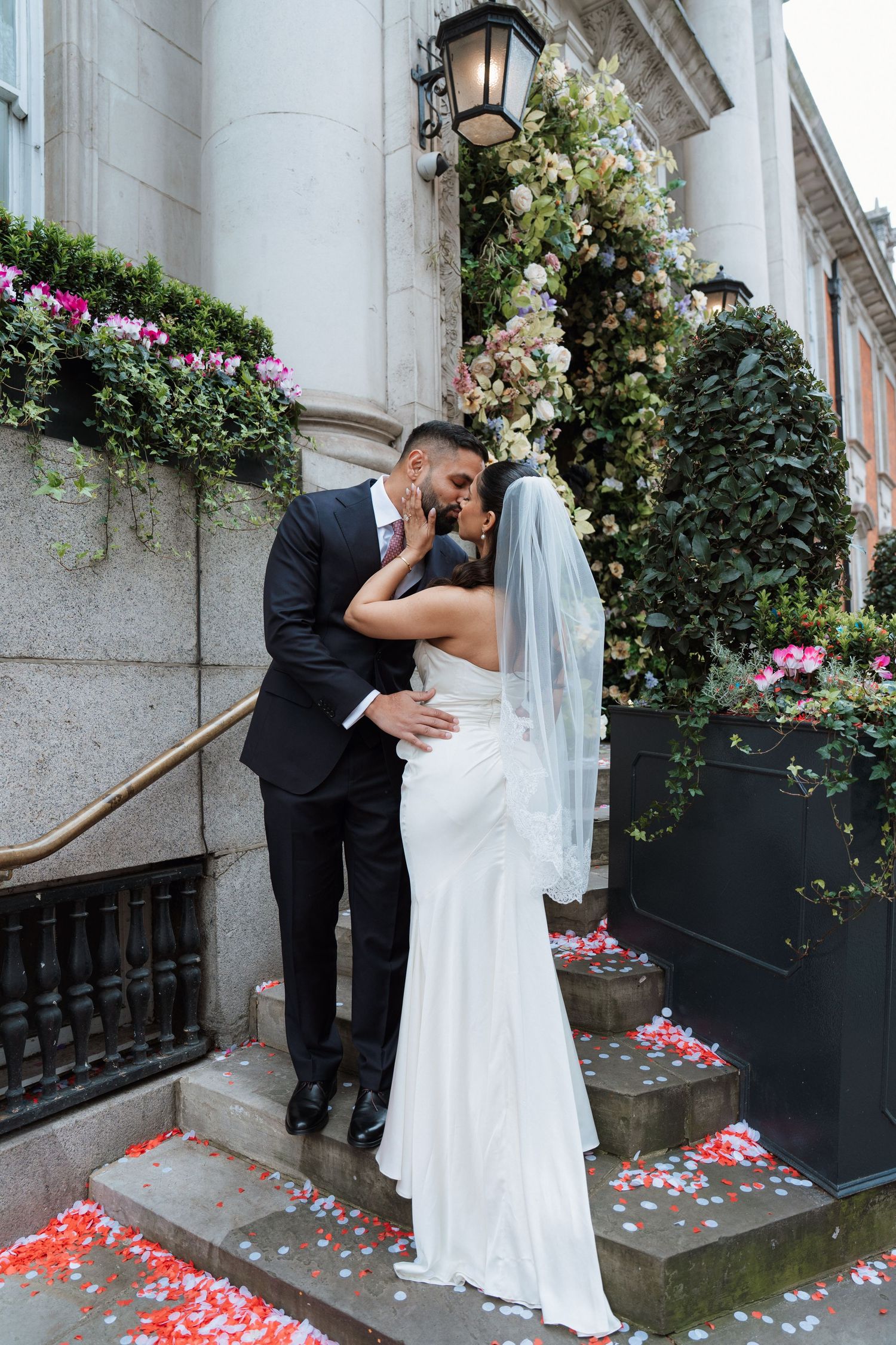 A romantic wedding couple shares an intimate moment on steps decorated with scattered rose petals and floral arrangements.