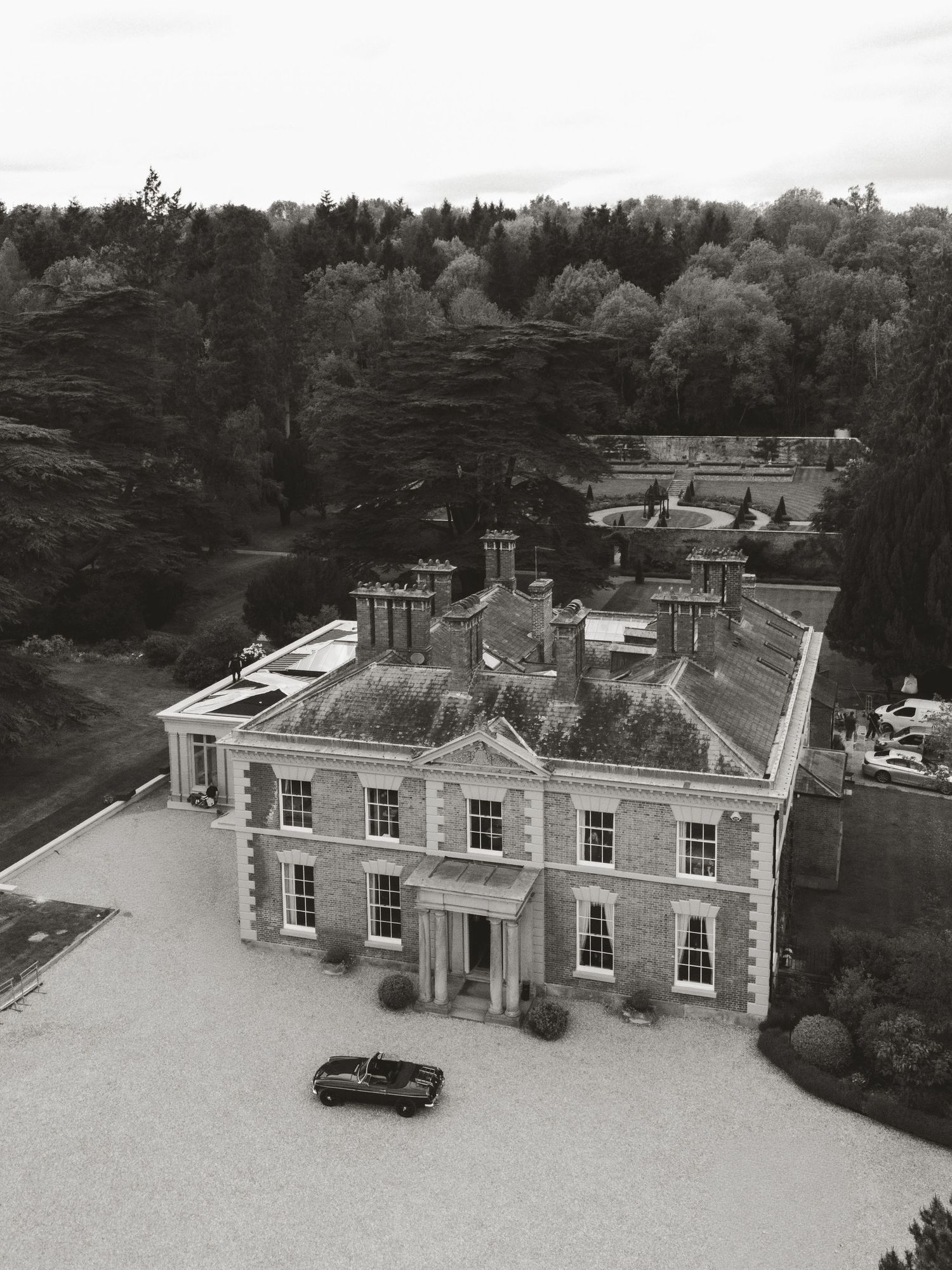 Aerial black and white photo of a grand English country manor house with formal gardens and a classic car parked outside.
