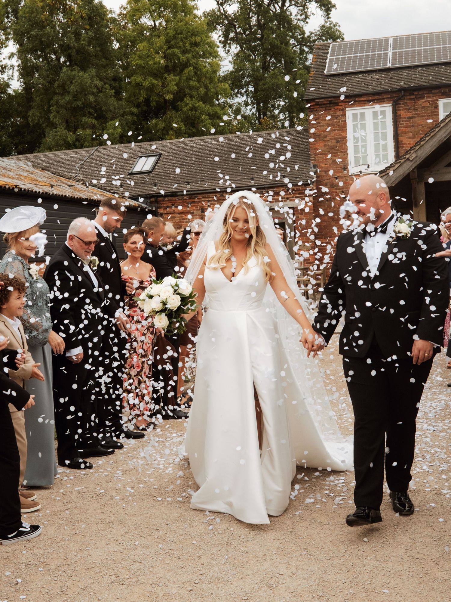 Wedding guests shower a couple with white confetti on a gravel path during a joyous outdoor celebration.