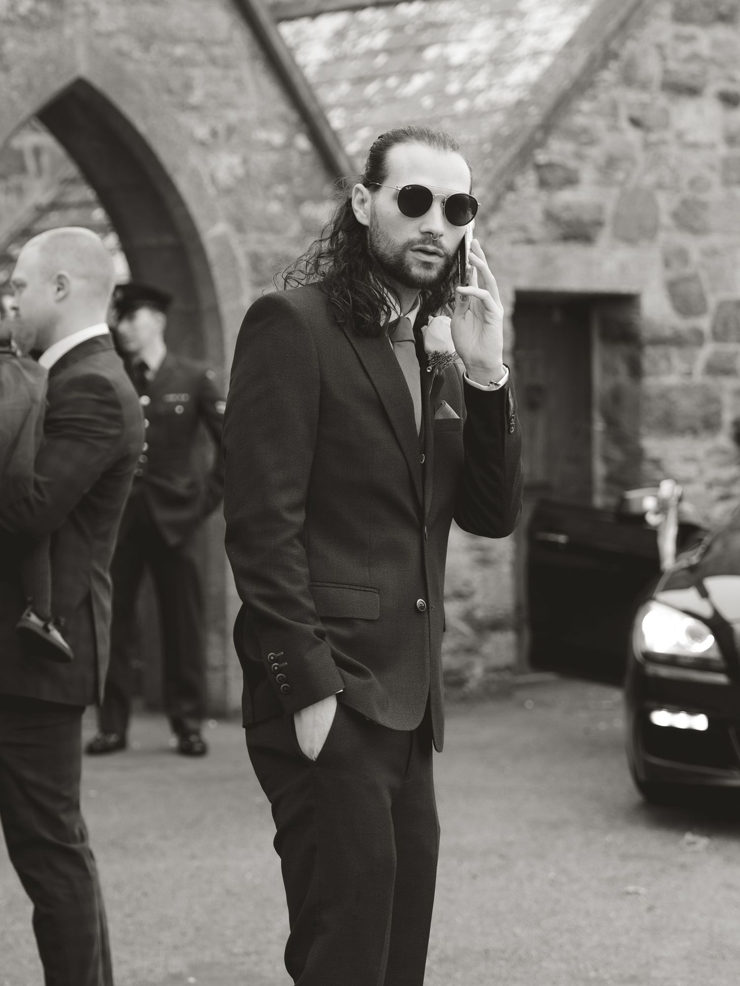 Men in black suits and sunglasses standing against stone wall backdrop.