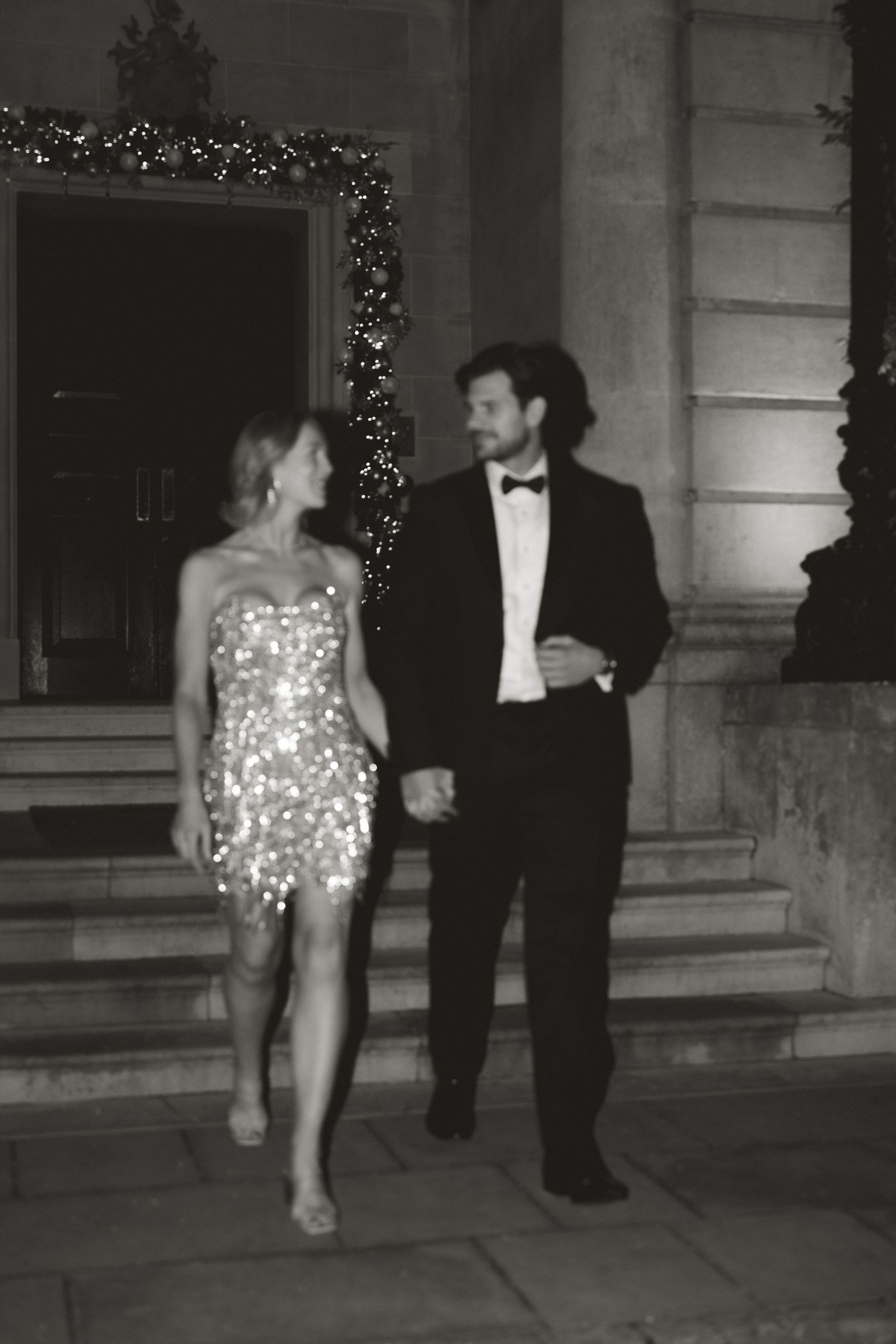 Elegant couple in formal attire, woman in sequin mini dress and man in tuxedo, posing outside decorated building.