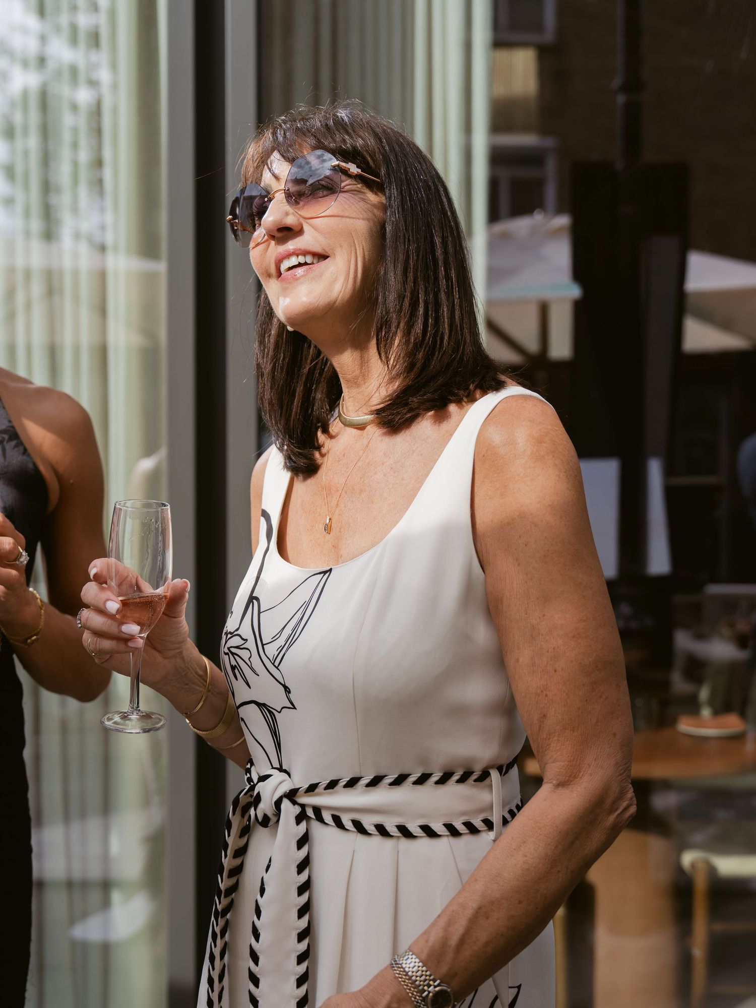 Woman in white floral dress and sunglasses smiling while holding champagne glass outdoors at social event.