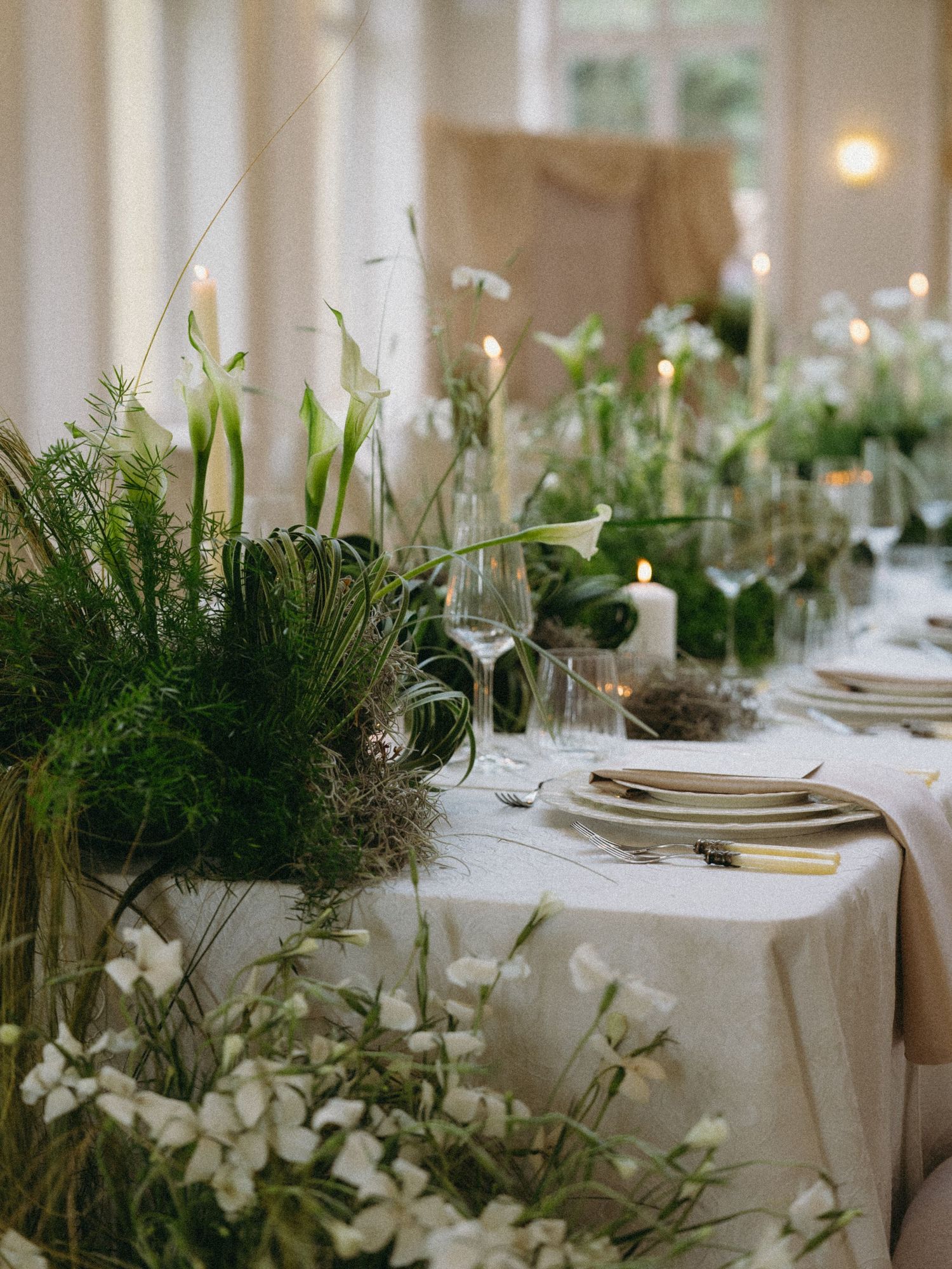 Elegant wedding table with white calla lilies, ferns, candles, and white linens for a lush garden-inspired reception.