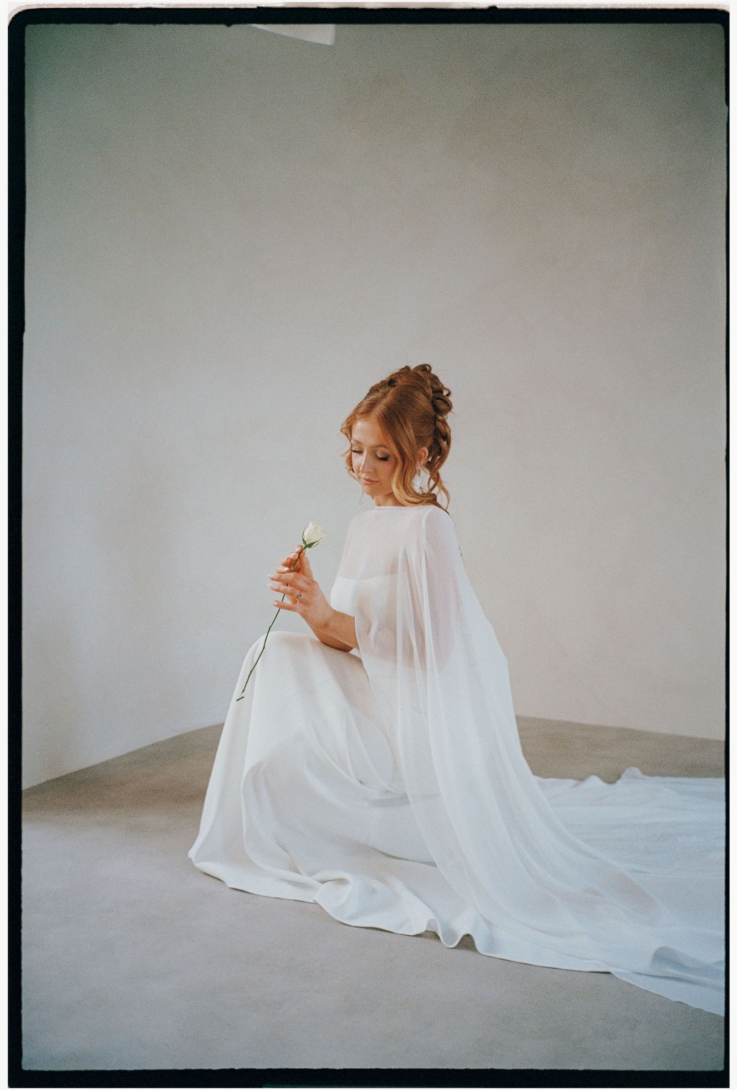 Bride in flowing white gown kneeling gracefully in minimalist setting.