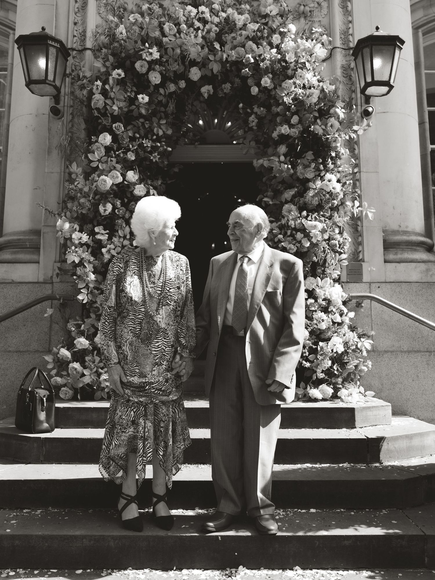 Elderly couple posing together on steps adorned with floral arch in black and white photo.