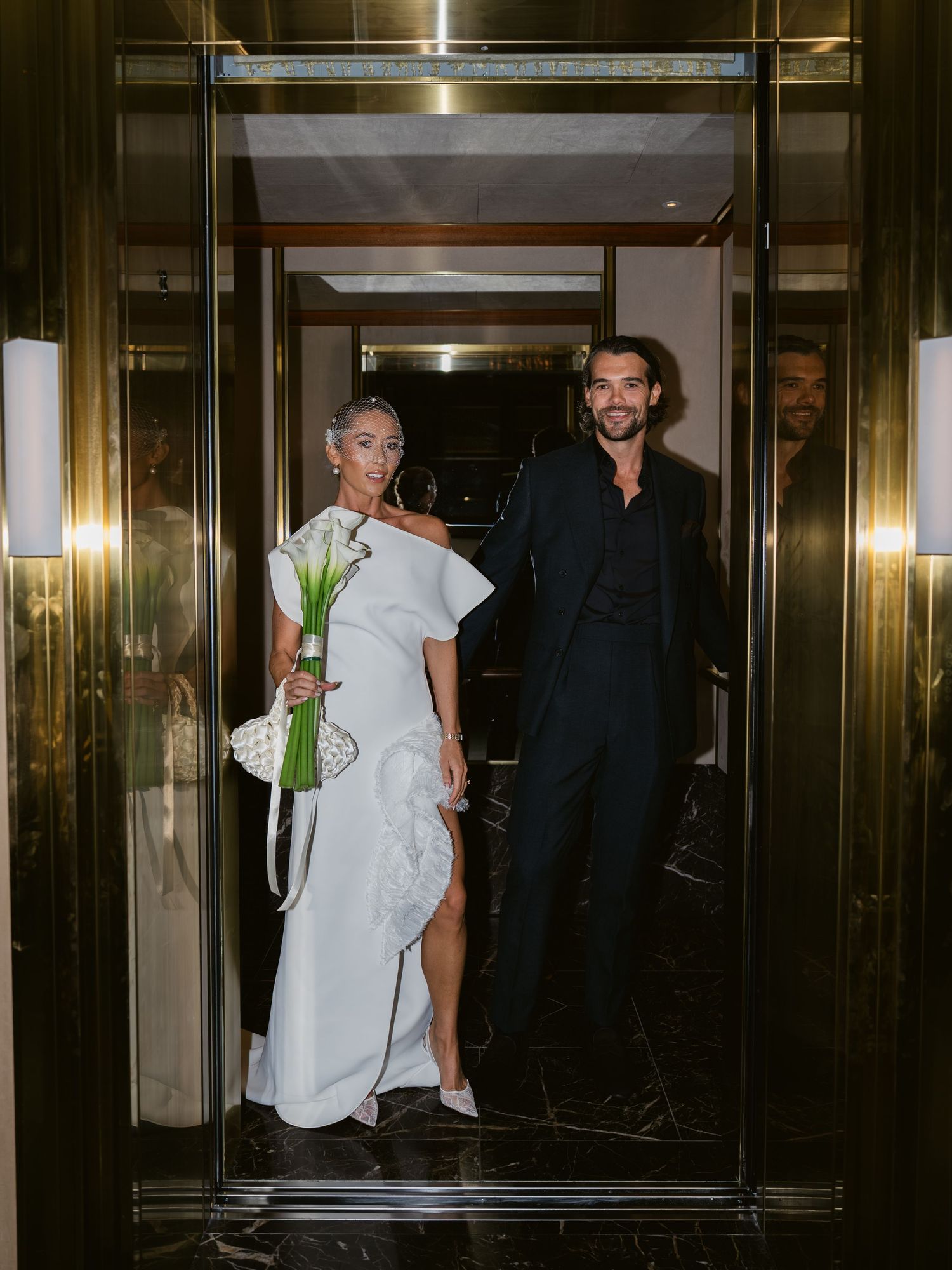 A couple in wedding attire poses in a gold-framed doorway, bride in white gown holding calla lilies, groom in navy suit.