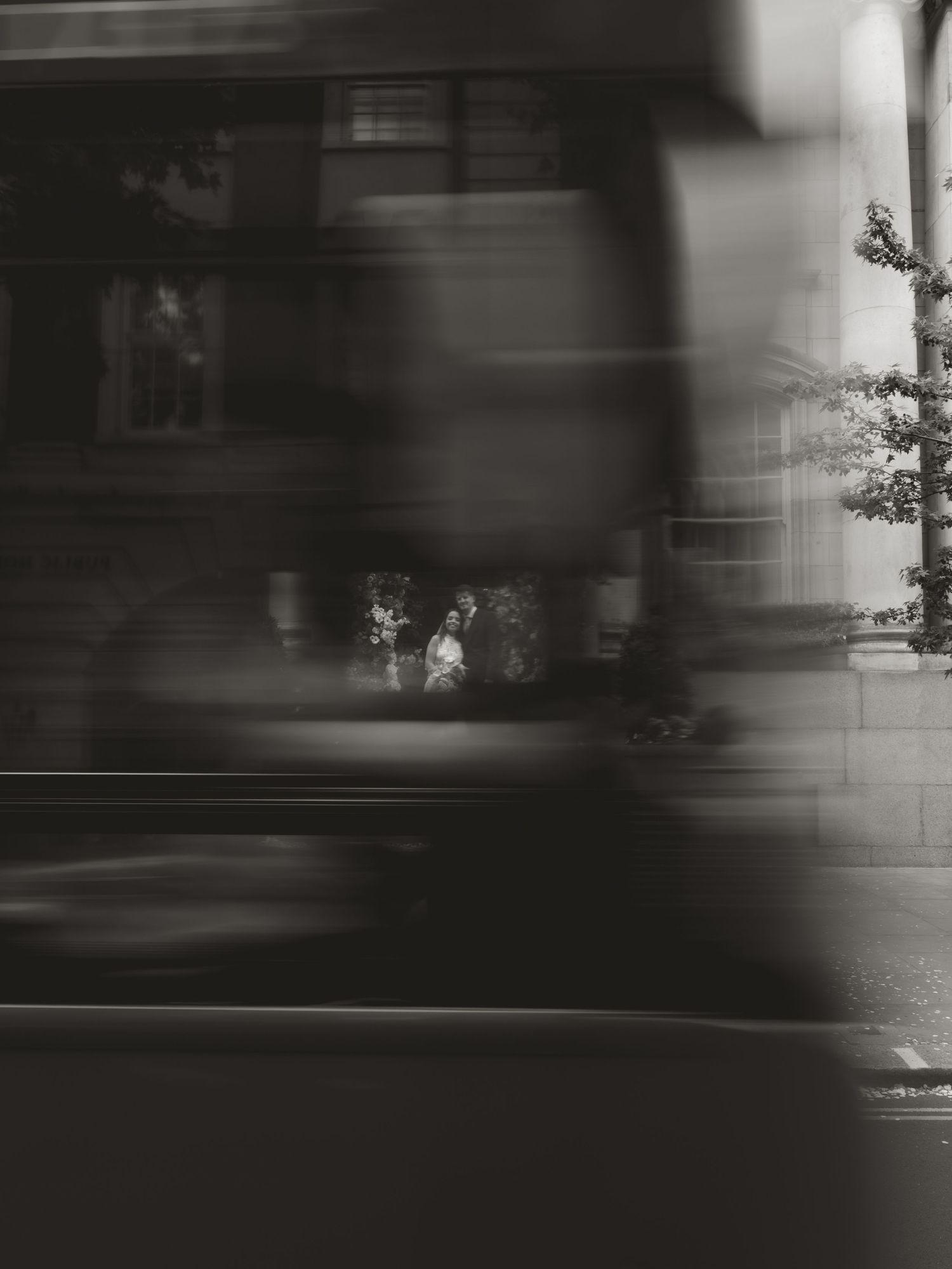Blurred black and white photo of a moving bus on a city street with pedestrians and trees in the background.