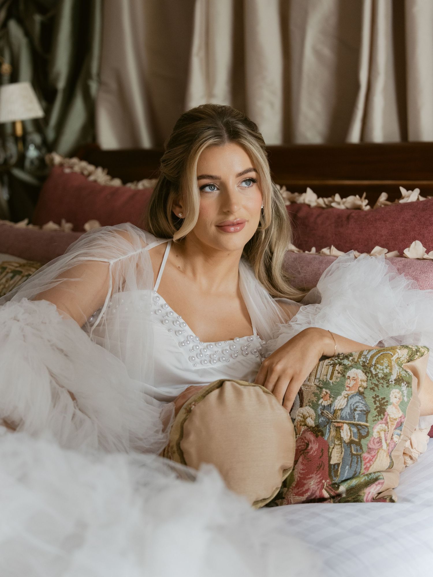 A bride in a white lace bodice and tulle robe reclines gracefully on a bed with floral pillows.