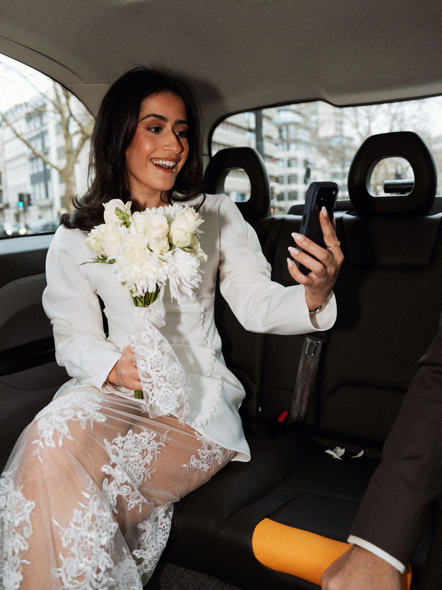Elegant lace wedding dress details captured in the back seat of a car with white floral bouquet.