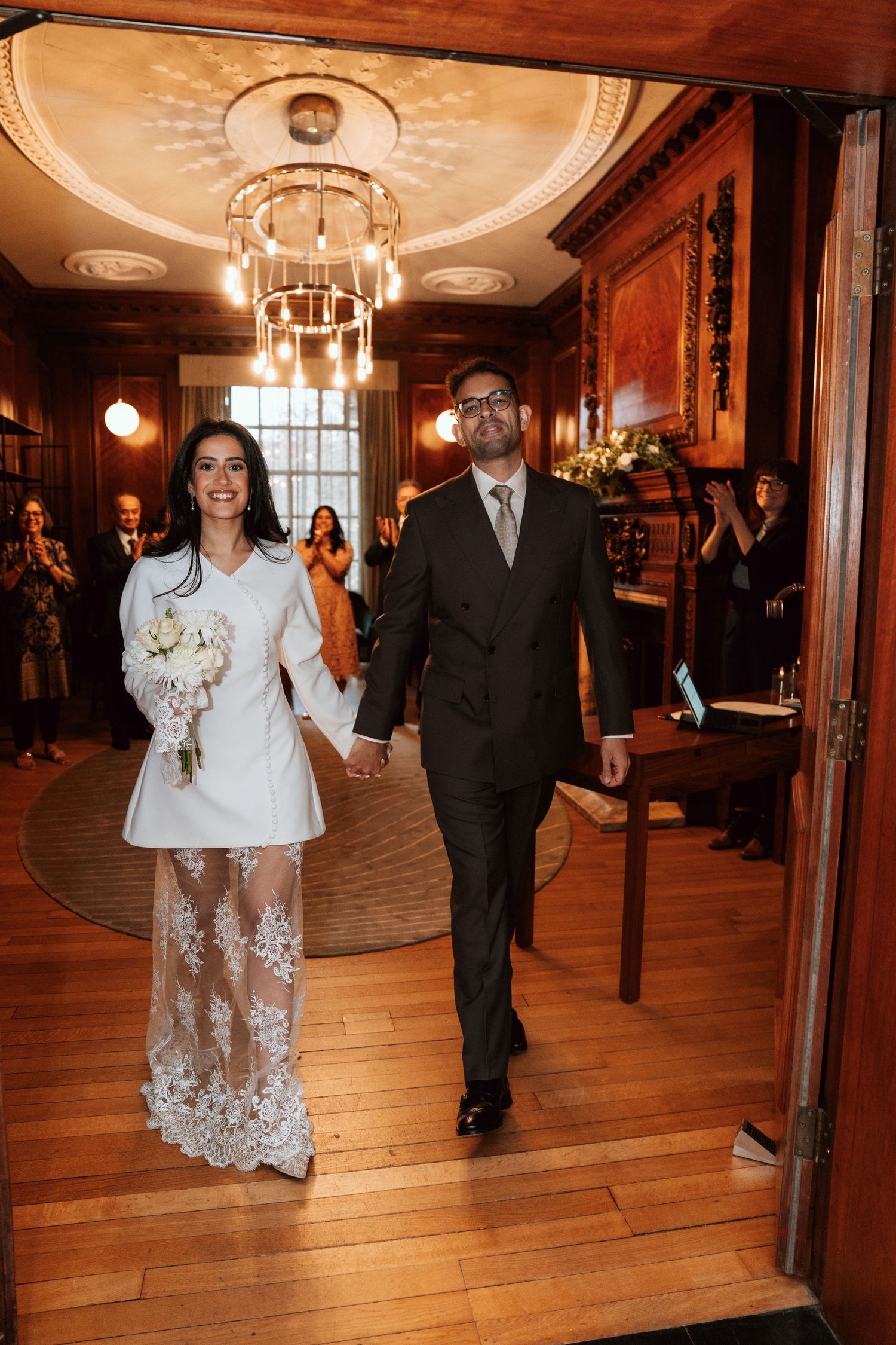Wedding couple walking through an elegant wood-paneled hallway with chandelier lighting.