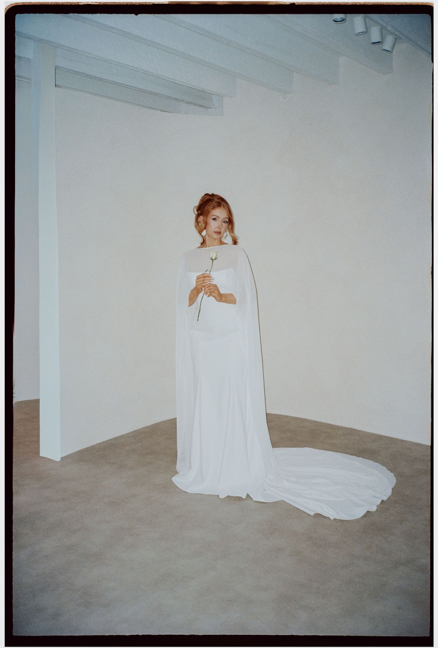 A bride in a flowing white cape-style wedding gown stands elegantly against a minimalist white wall.
