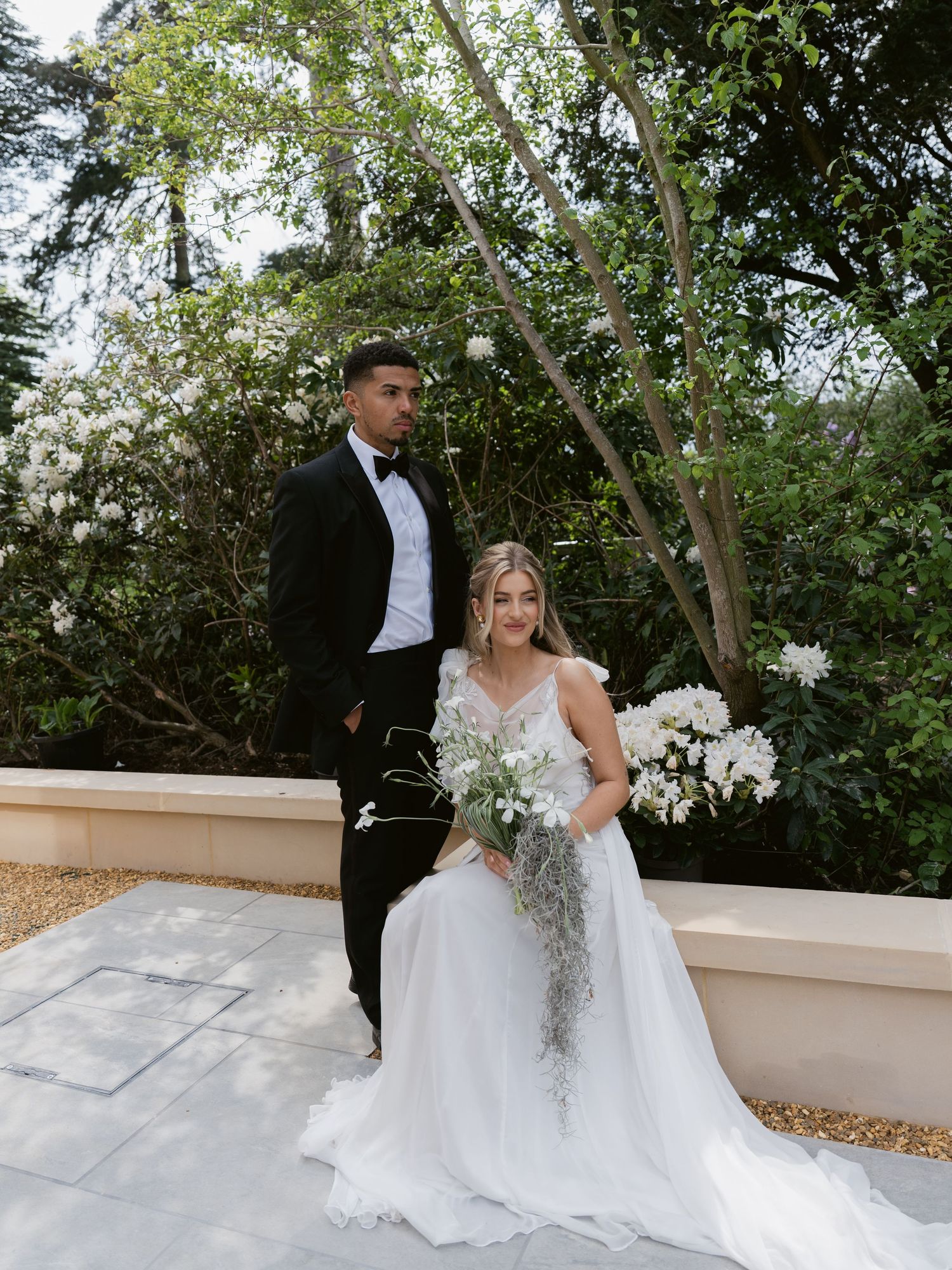 Bride in white gown holding floral bouquet sits beside groom in black tuxedo, posing outdoors with lush greenery backdrop.